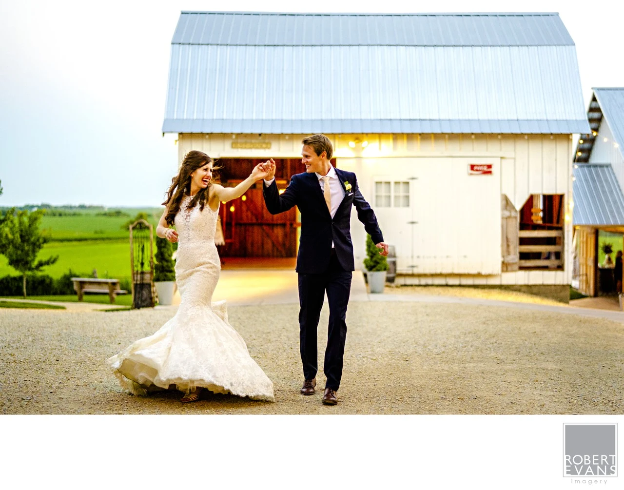 A groom spins his bride outside a white barn in warm evening light.