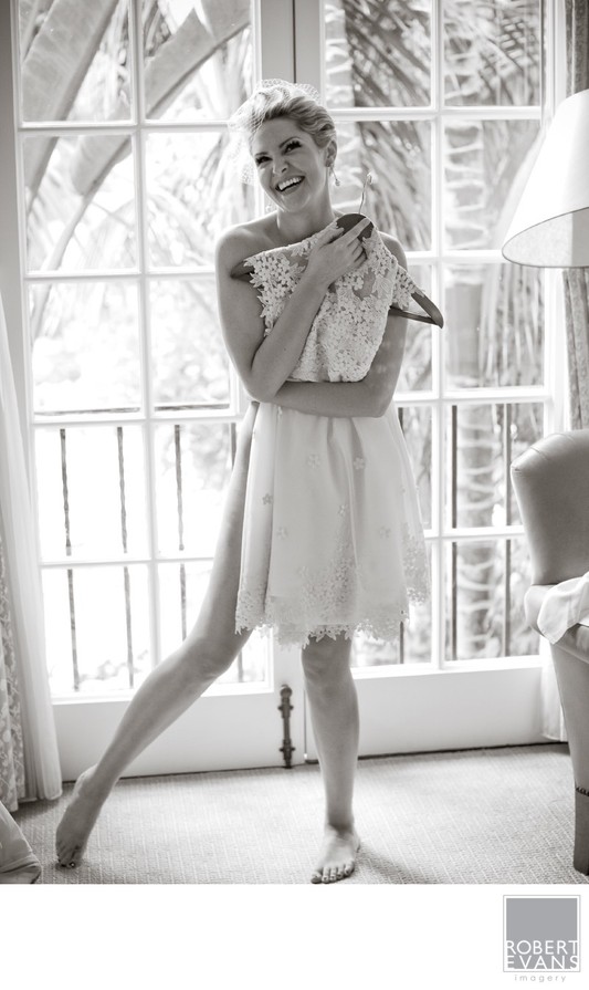A woman in a light robe stands beside bright windows holding a small item near her face.