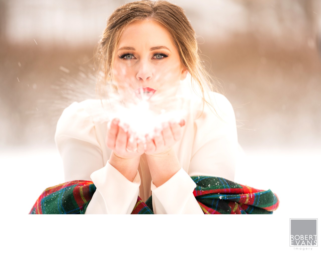 A bride blows snow from her hands toward the camera in soft winter light.