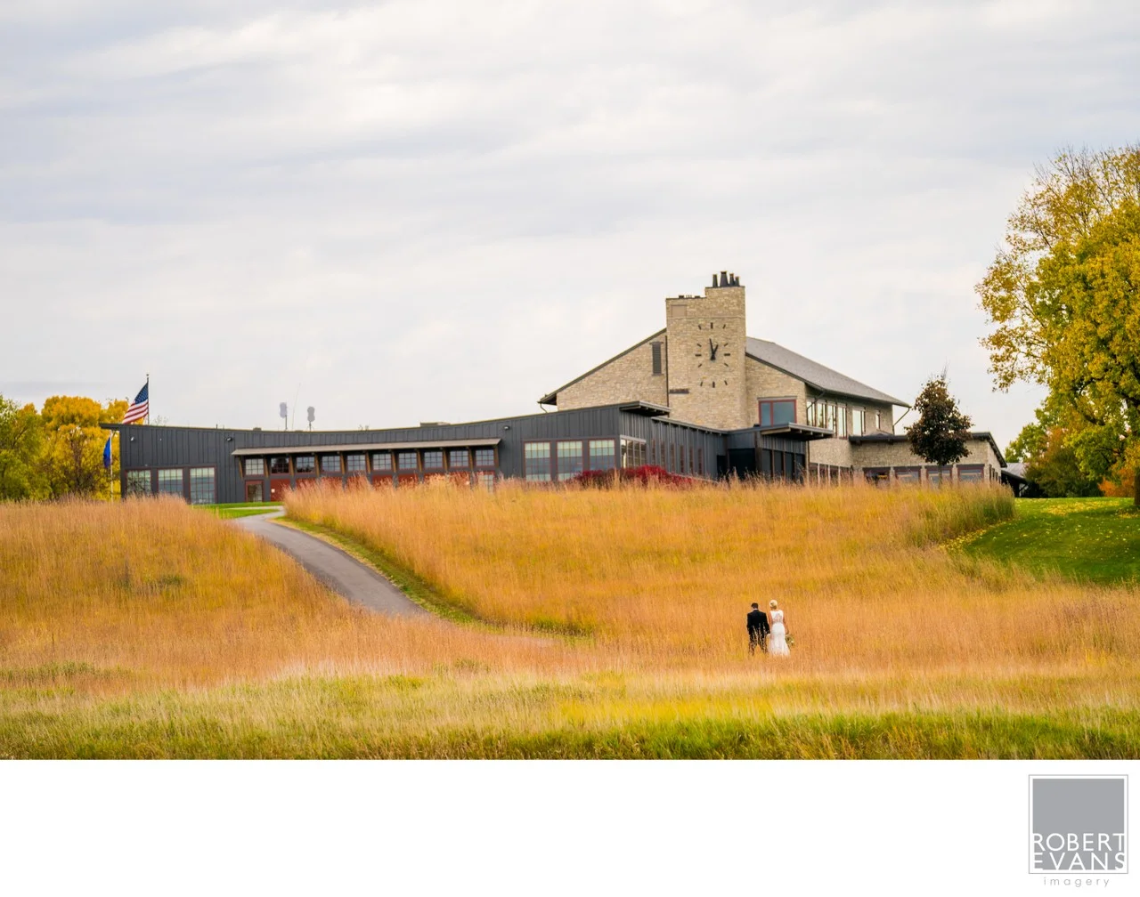 A couple walks through a broad golden meadow below a large clubhouse.