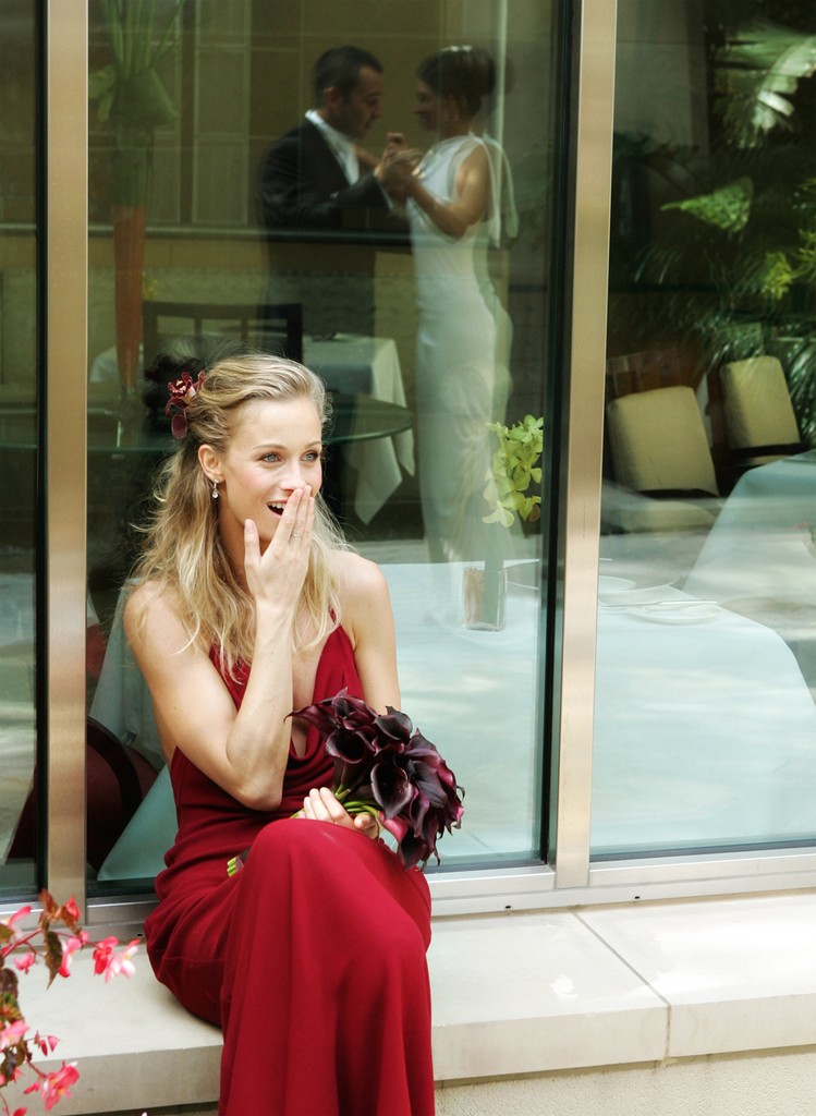 A woman in a red dress reacts while a wedding couple is reflected in glass behind her.