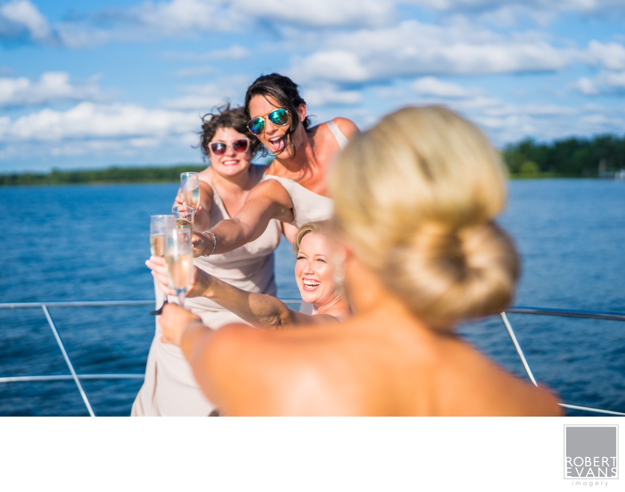 Smiling wedding guests gather beside a bride on a boat under a bright blue sky.