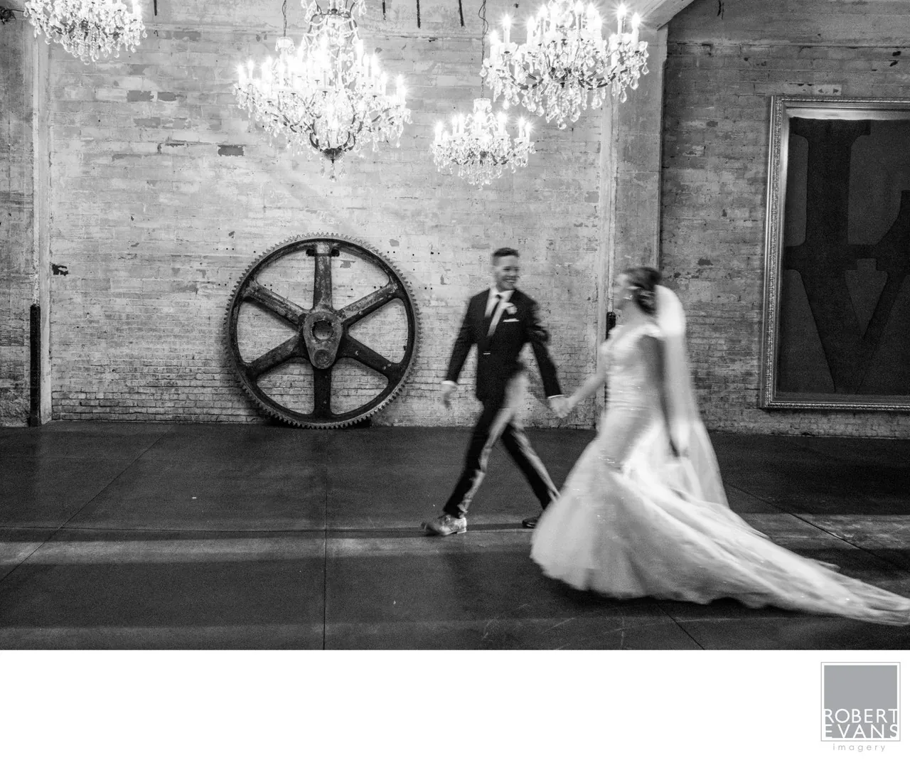 A groom turns toward a bride approaching beneath chandeliers in an industrial wedding venue.