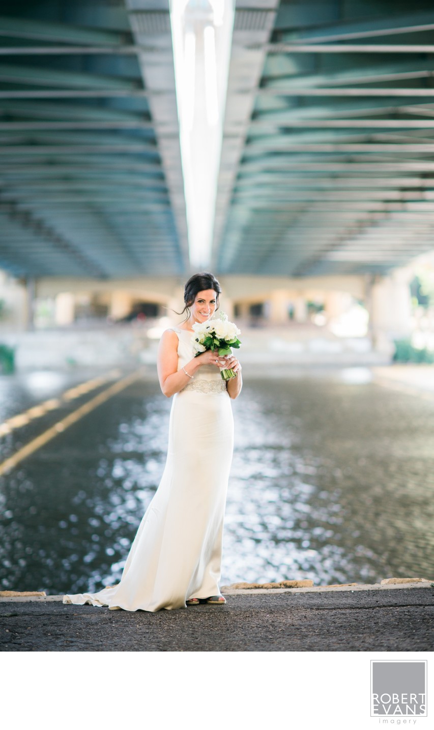 A bride holding flowers stands beside water beneath the geometric beams of a bridge.