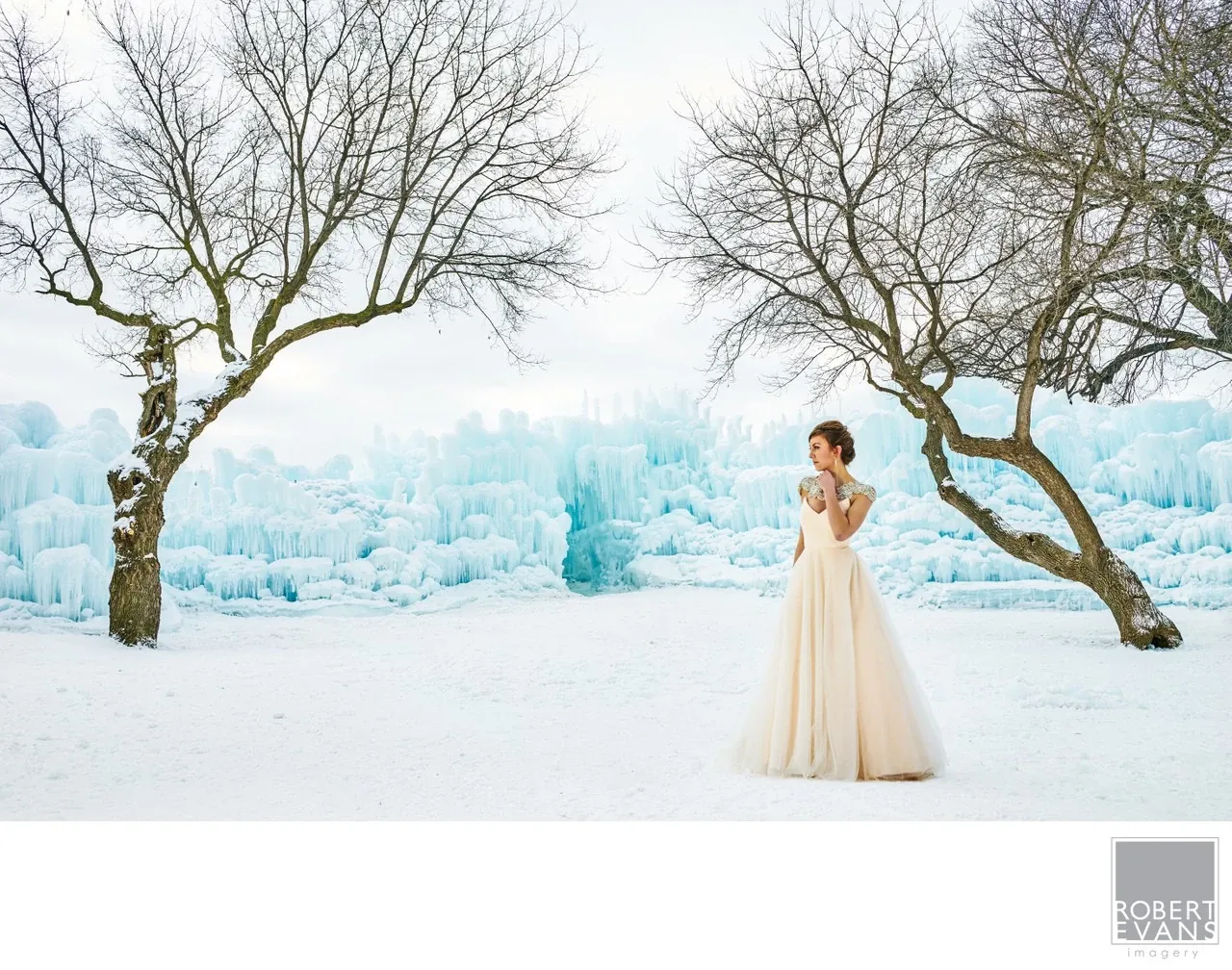 A bride stands between bare trees with blue ice formations behind her in a snowy landscape.