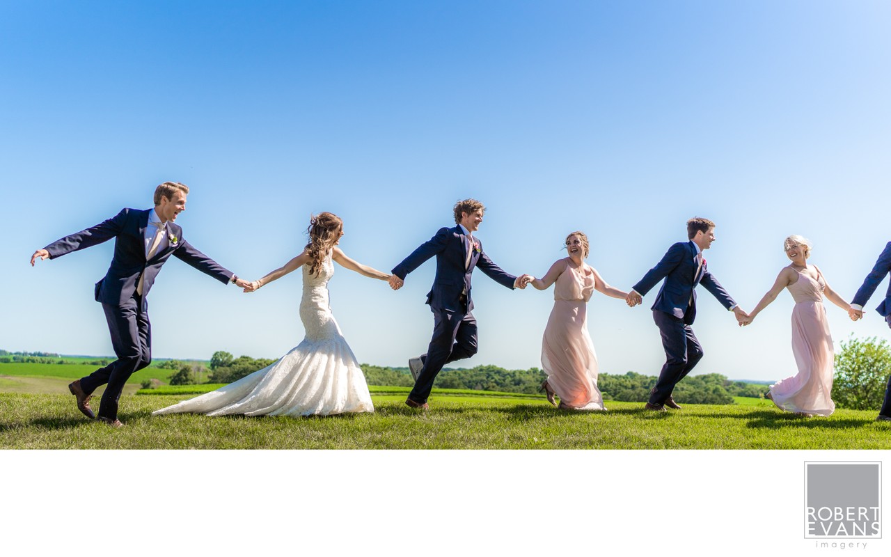 A wedding party runs hand in hand across a grassy hill beneath a clear blue sky.