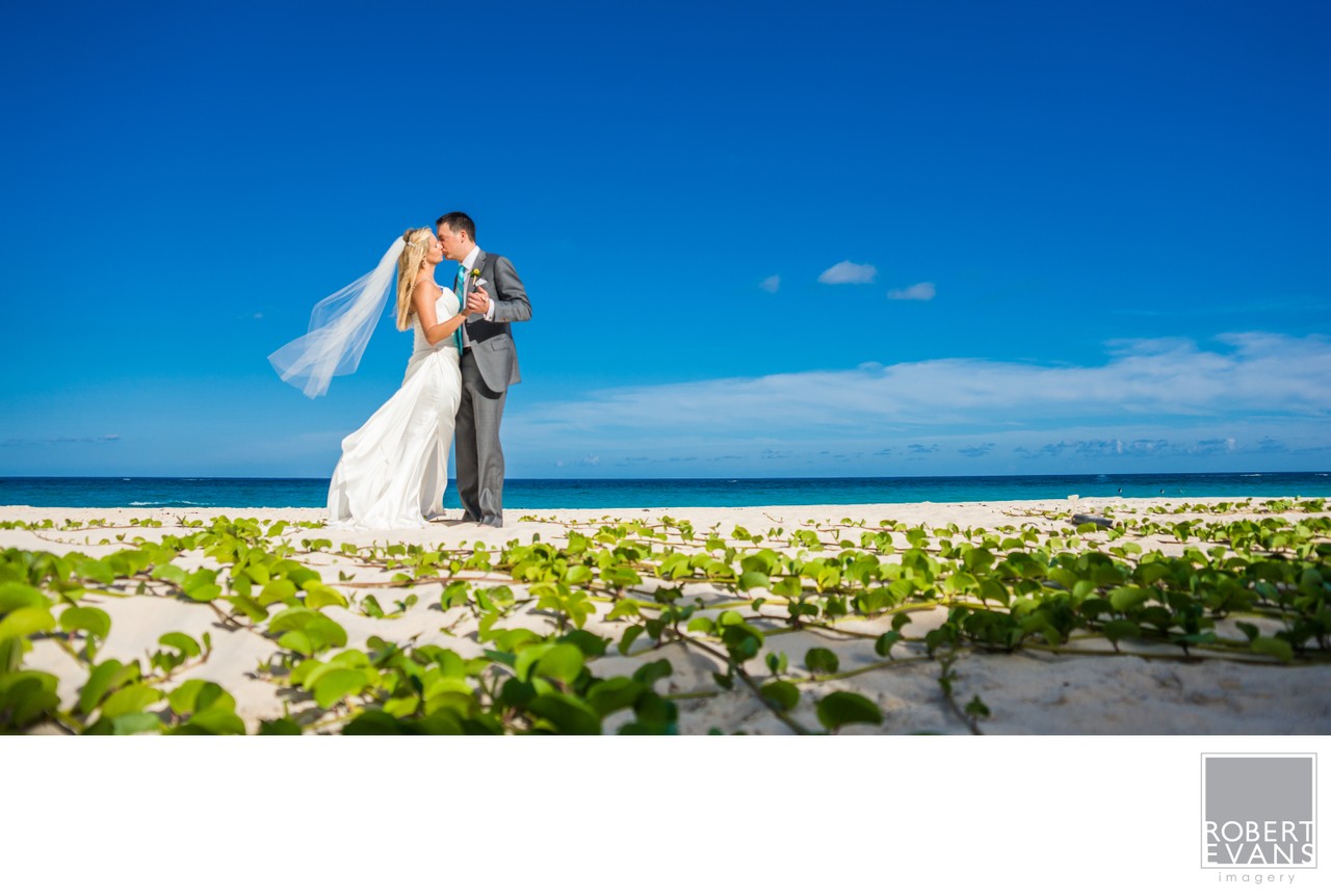 A bride and groom embrace on a sandy beach beneath a vivid blue sky.