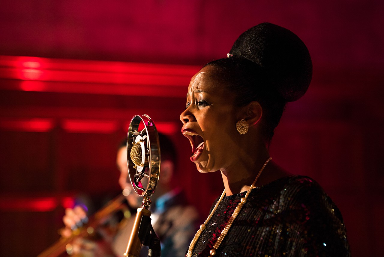 A singer performs into a vintage-style microphone under warm red stage lighting.
