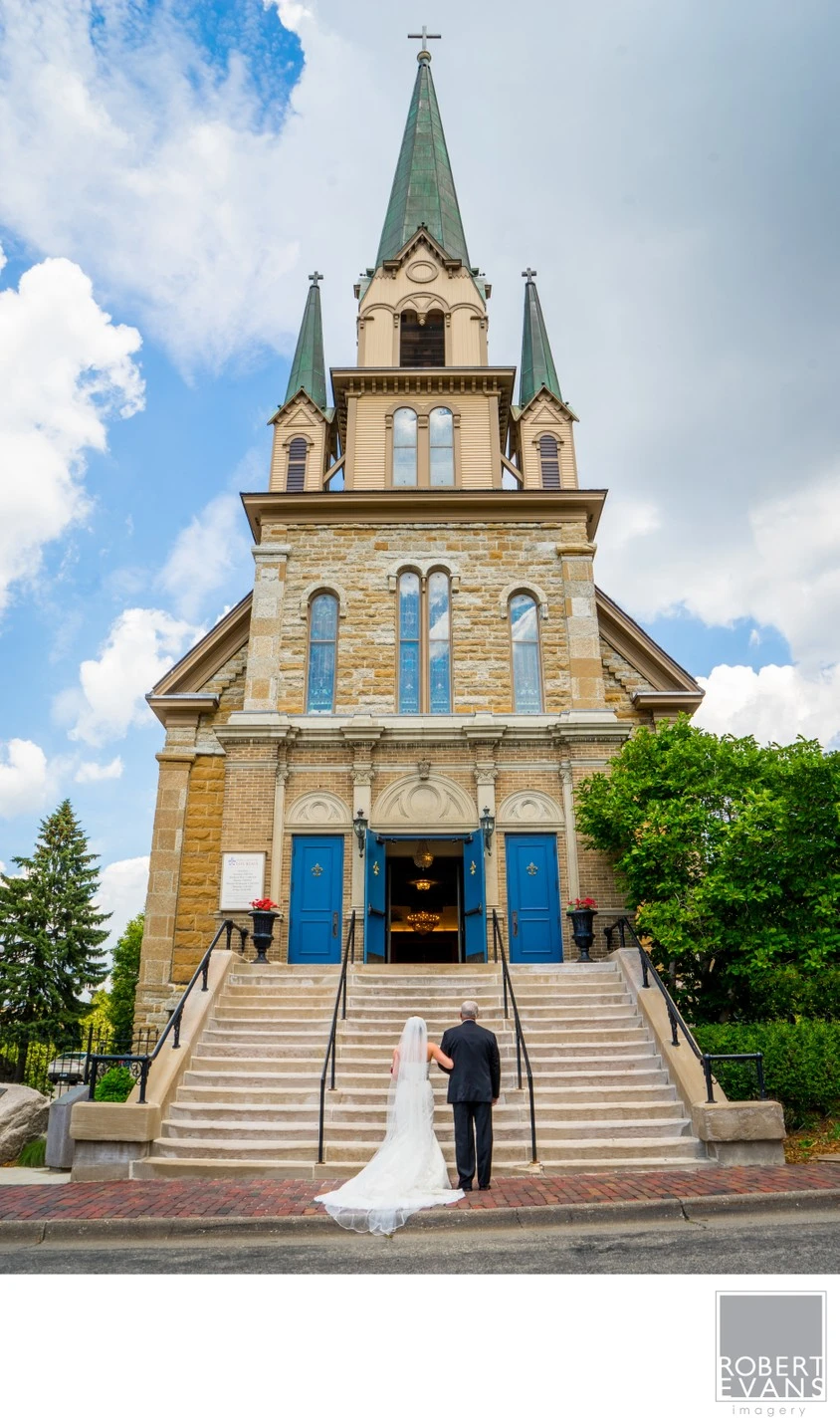 A bride and her father wait on the steps beneath a tall church steeple.