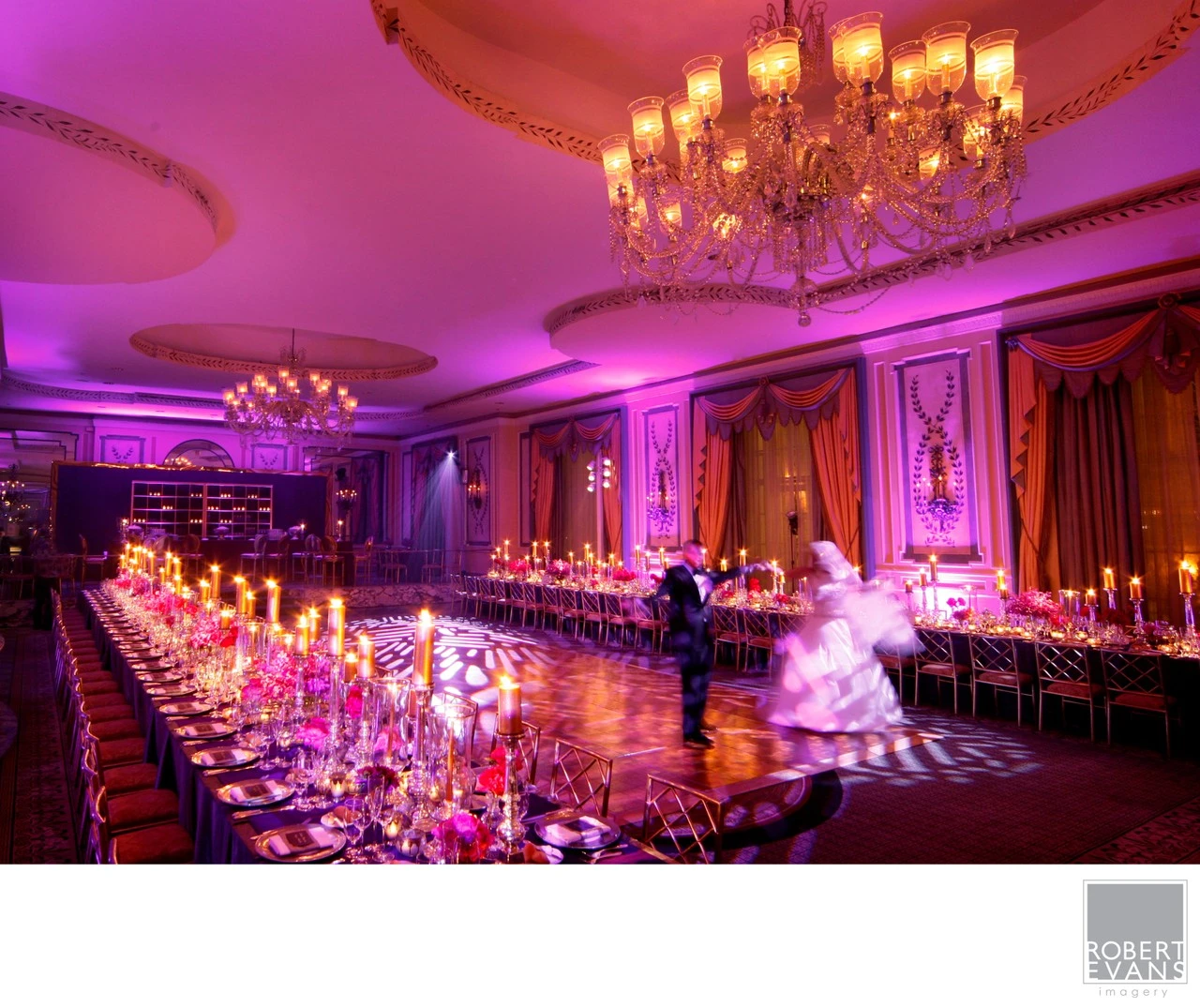 A bride and groom dance in a grand ballroom illuminated with candles and pink light.