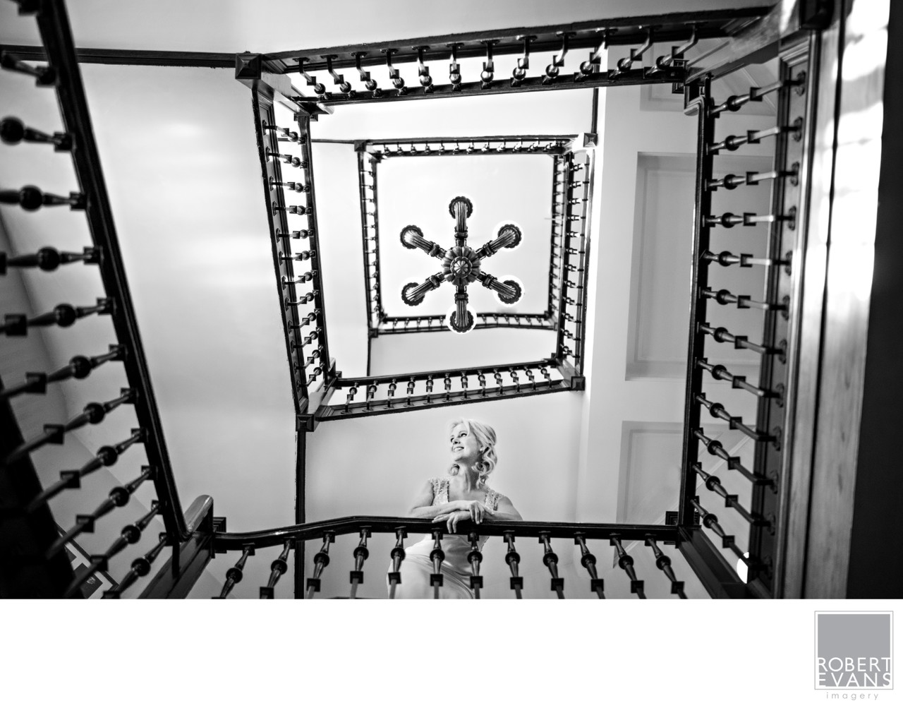 A bride is framed by a geometric stairwell and chandelier photographed from below in black and white.