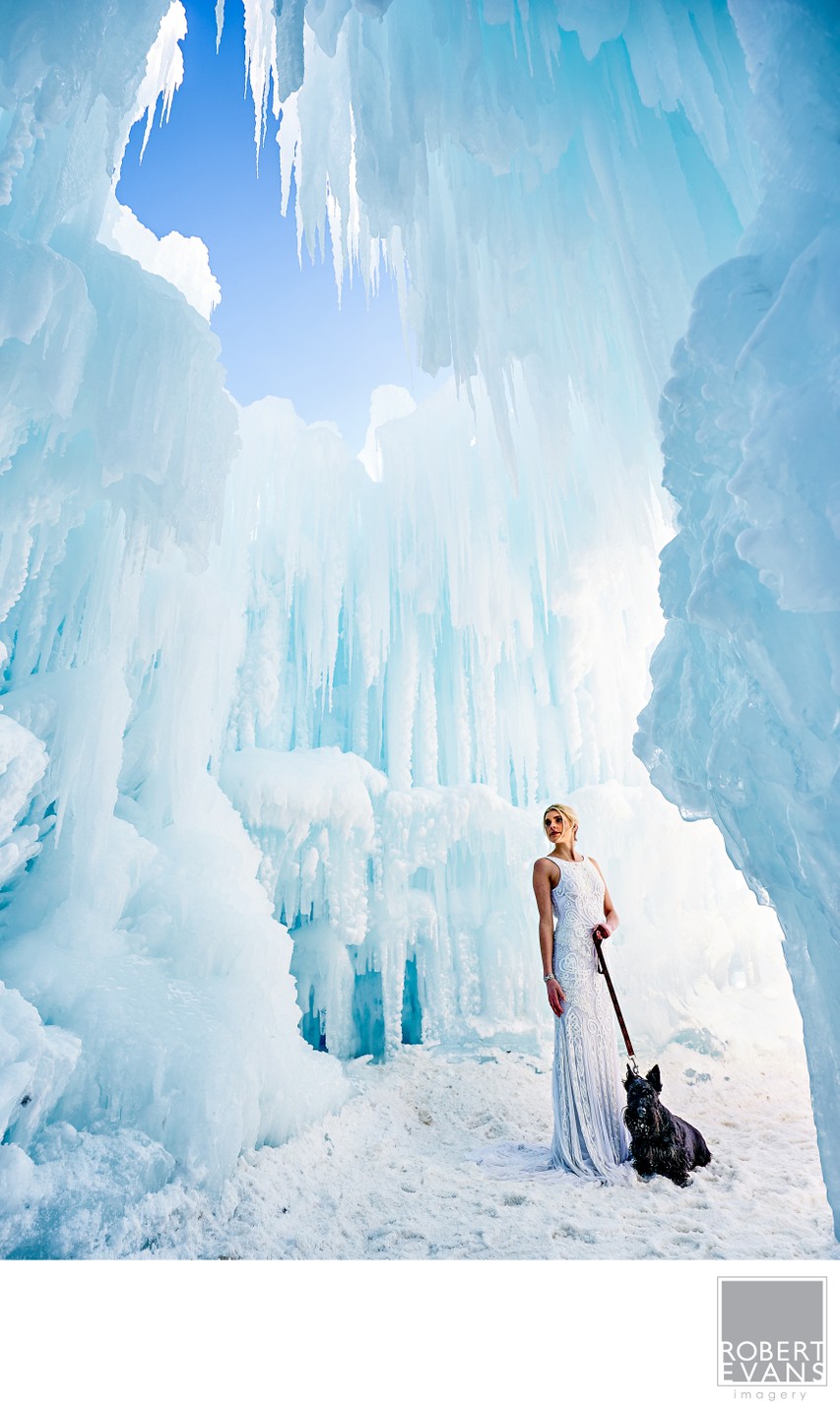 A bride stands beneath tall blue ice formations with a dark bouquet at her side.