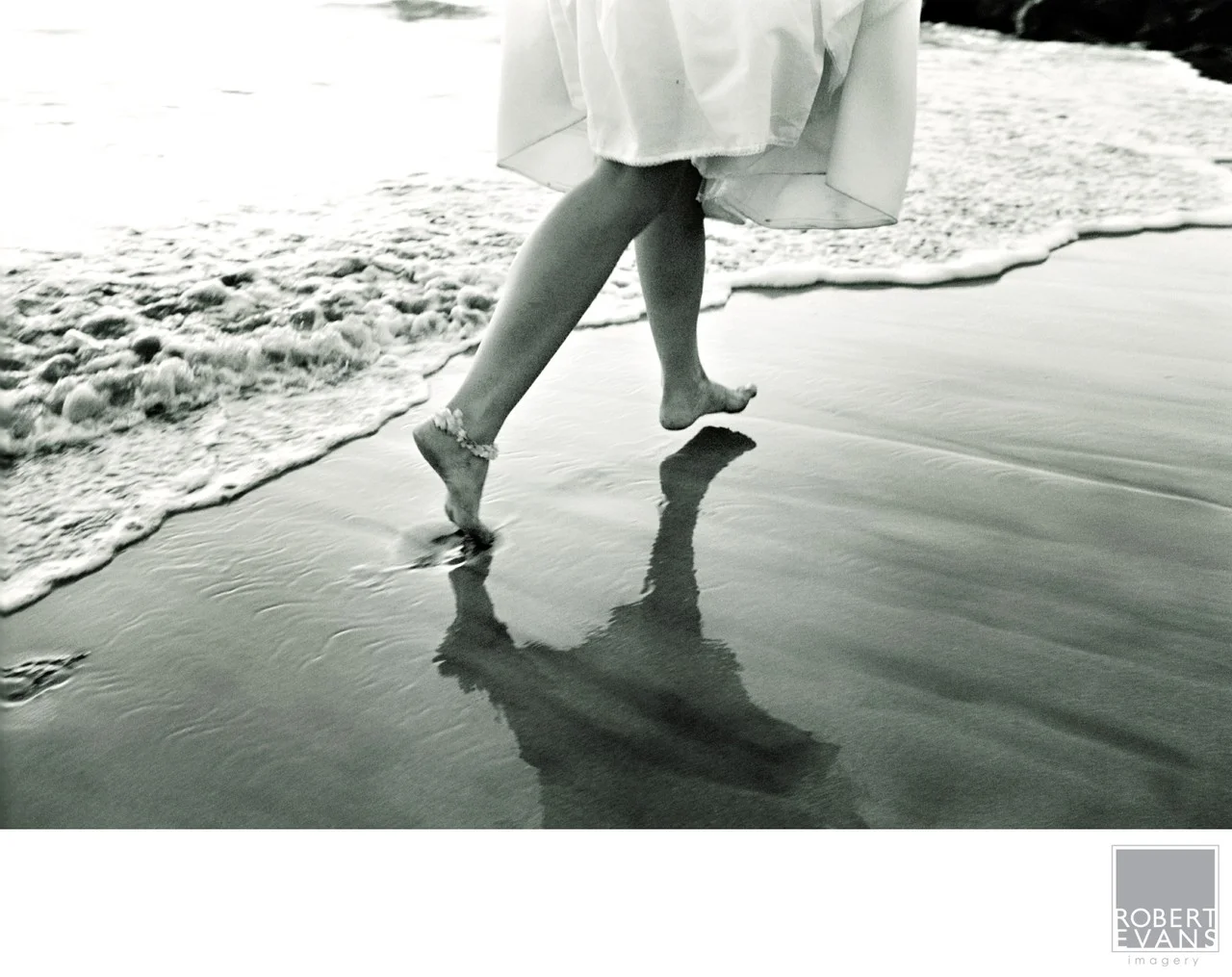 A bride’s bare feet and flowing skirt are reflected in wet sand beside gentle waves.