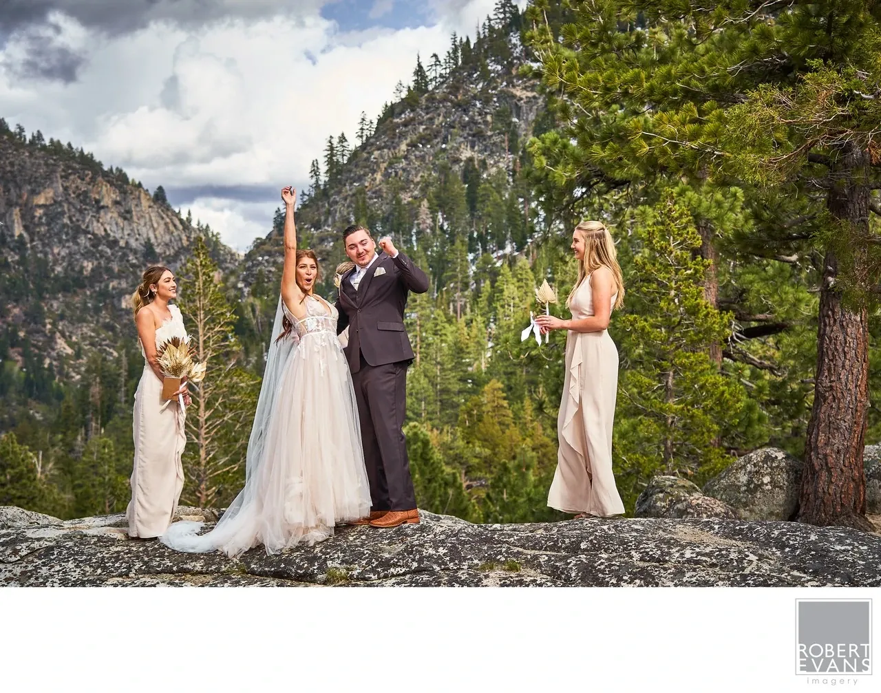 A newly married couple raises their arms beside attendants on a rocky overlook above forested mountains.