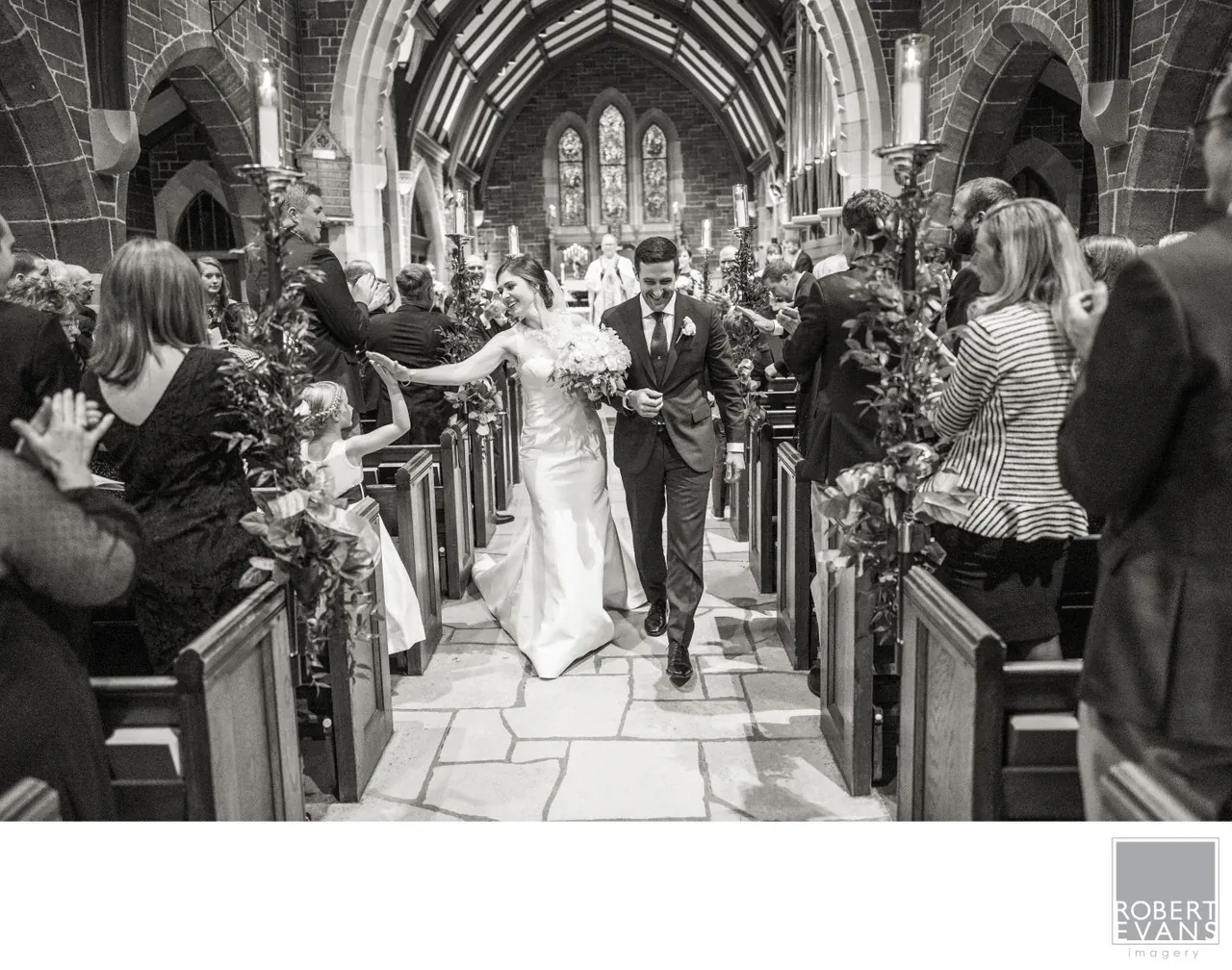 A bride and groom walk together down the aisle as guests applaud in black and white.
