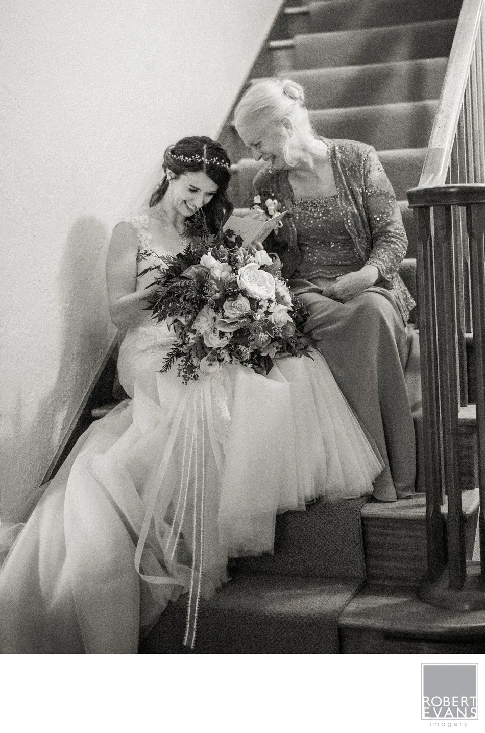 A bride and another woman sit closely together on a staircase in black and white.
