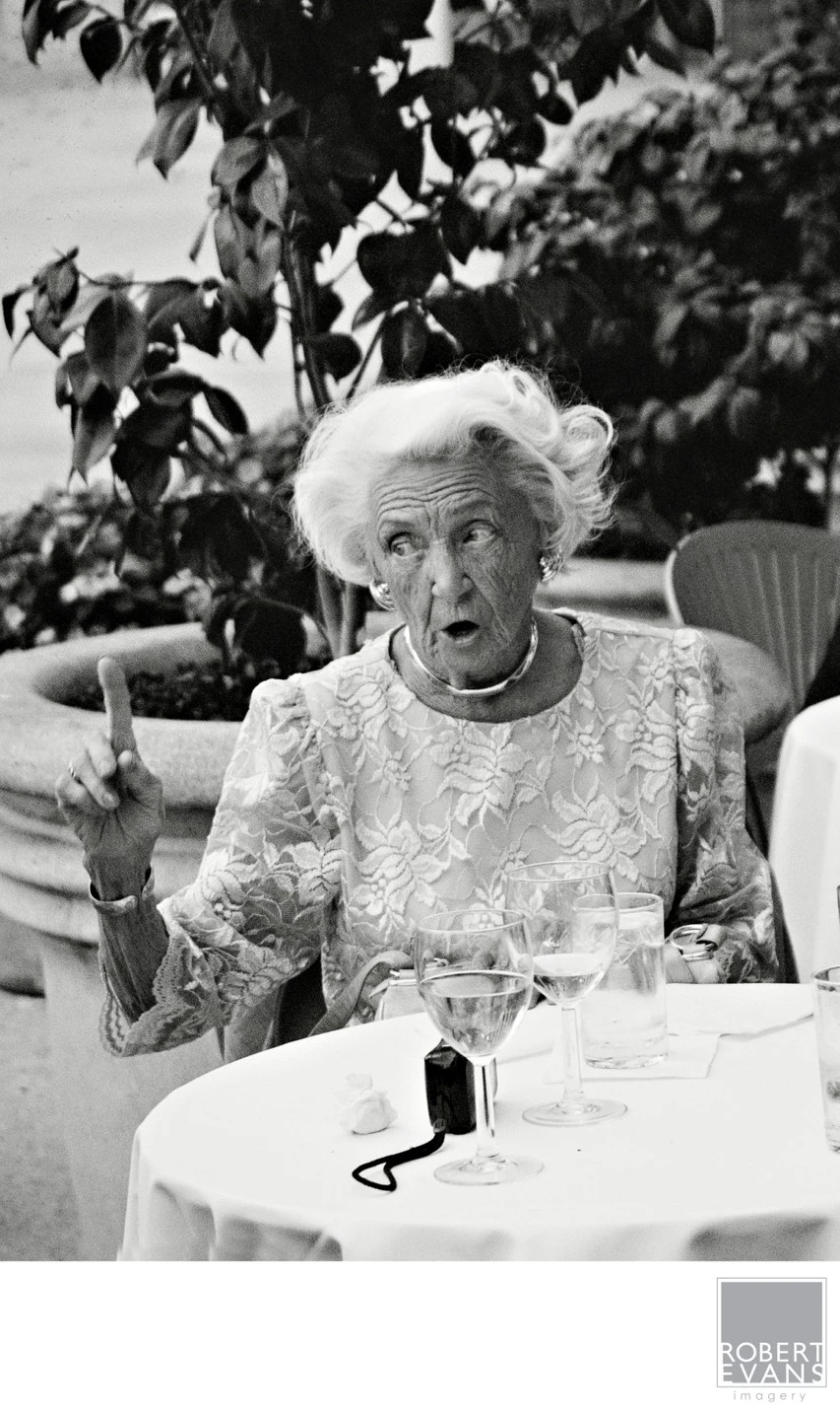An older woman gestures with a raised finger while seated among wedding guests in black and white.