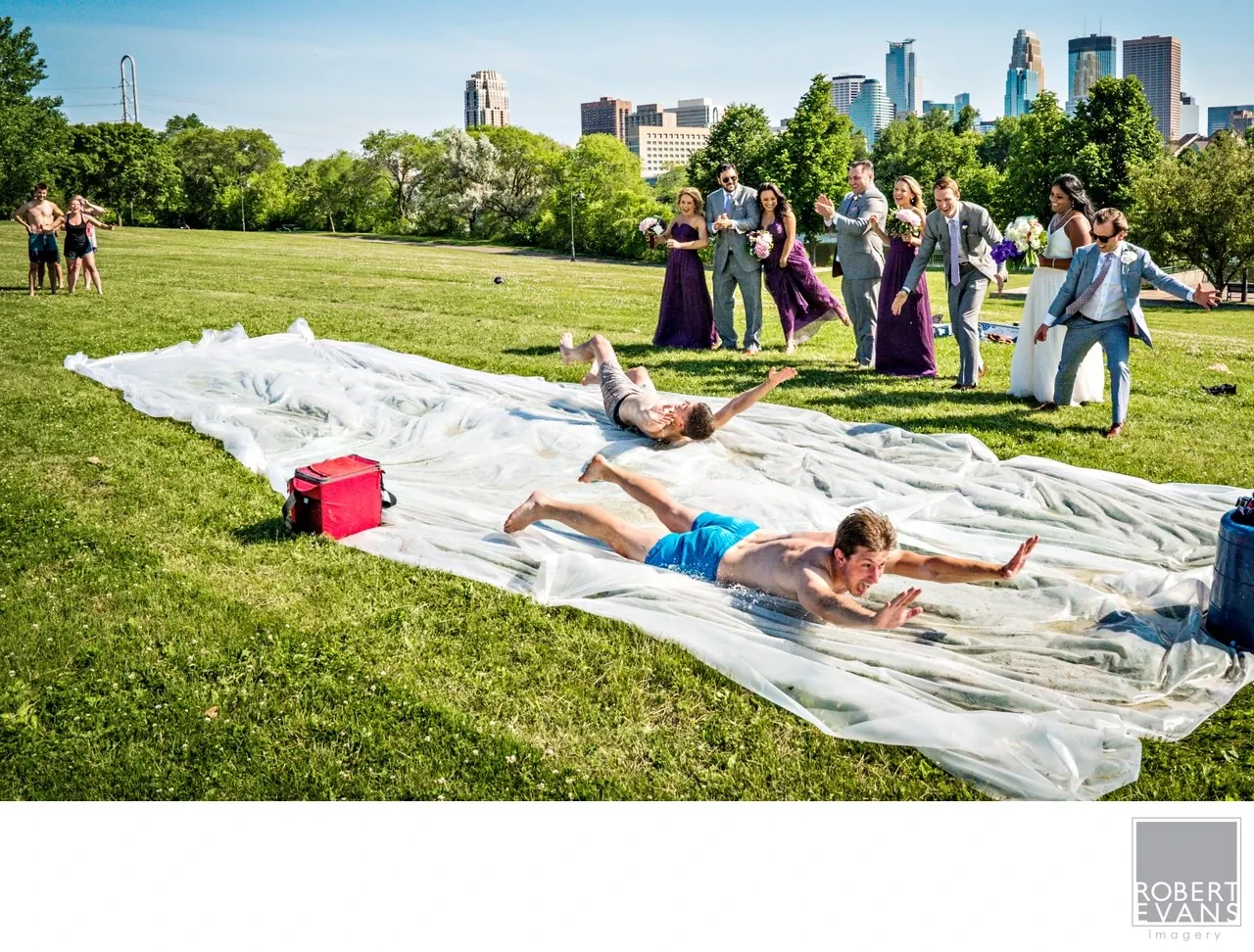 Wedding guests slide across a plastic water slide on grass while the wedding party watches.