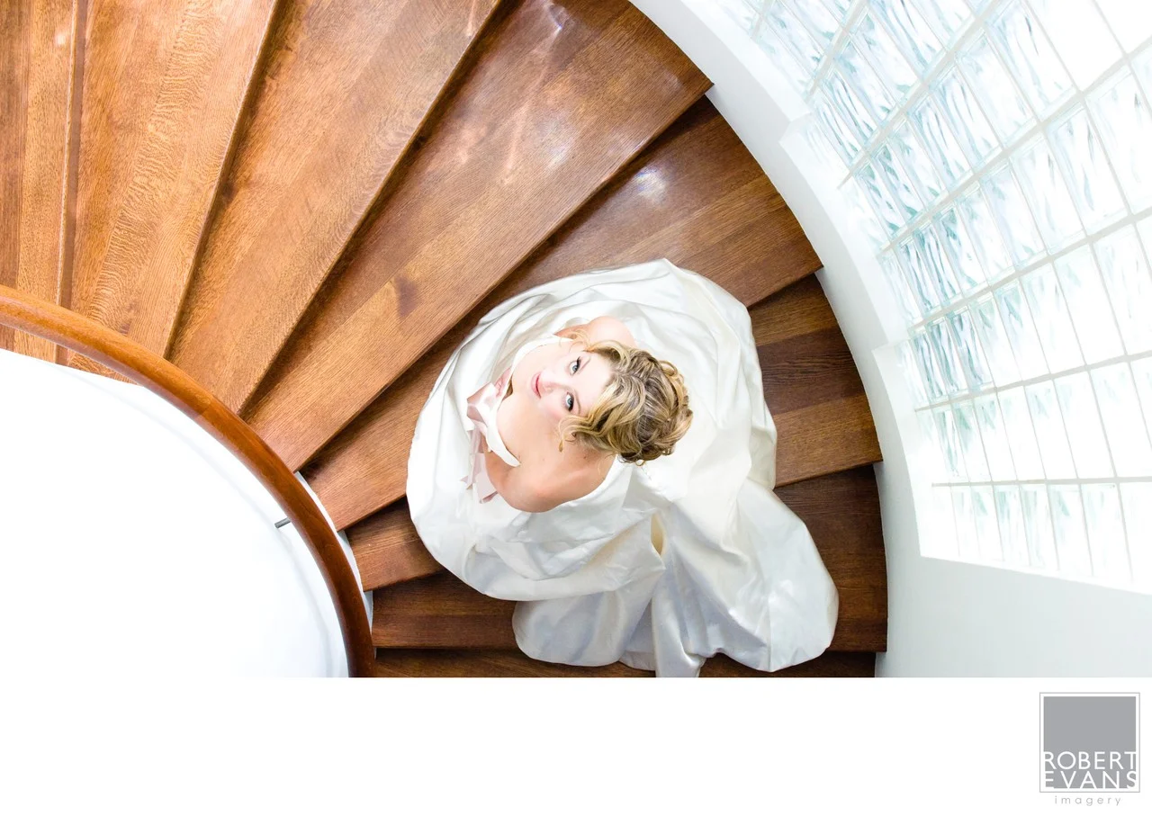 A bride looks upward from a curved wooden staircase beside glass-block windows.
