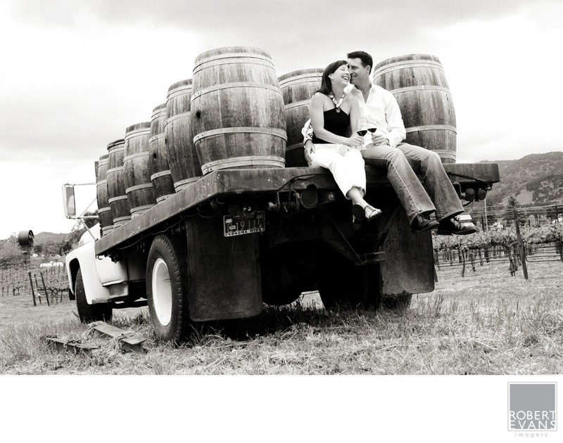 A couple kisses while seated on a vintage truck loaded with barrels in black and white.