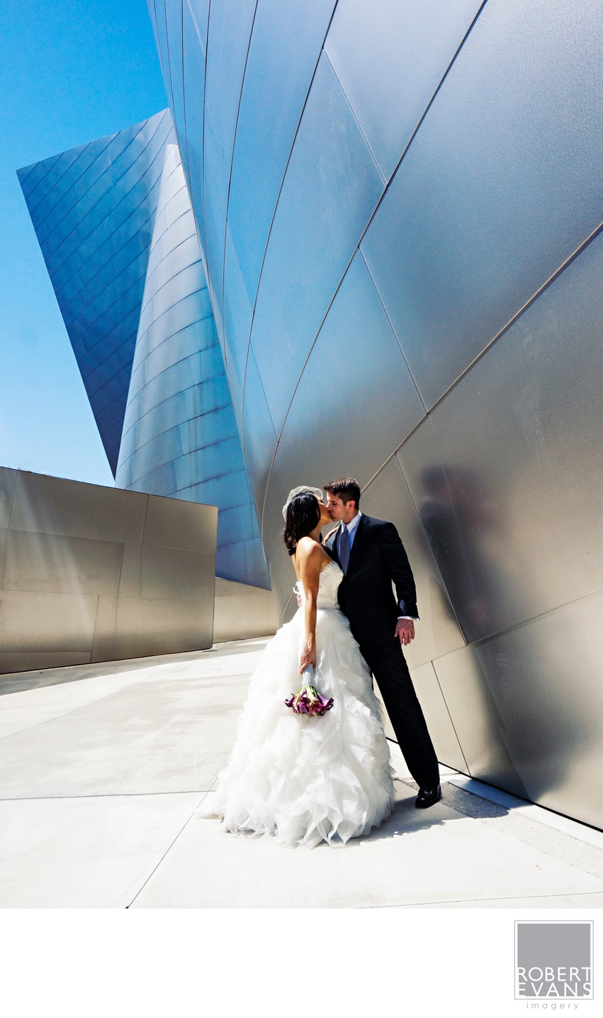 A bride and groom lean together beside sculptural metal walls in bright daylight.
