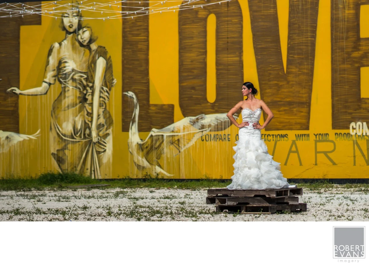 A bride extends her veil beside a large gold-toned mural on an urban wall.