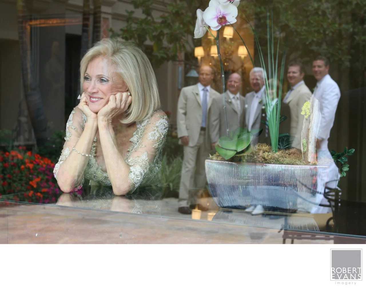 A bride rests her chin on her hands as reflections of people and greenery overlap the window.