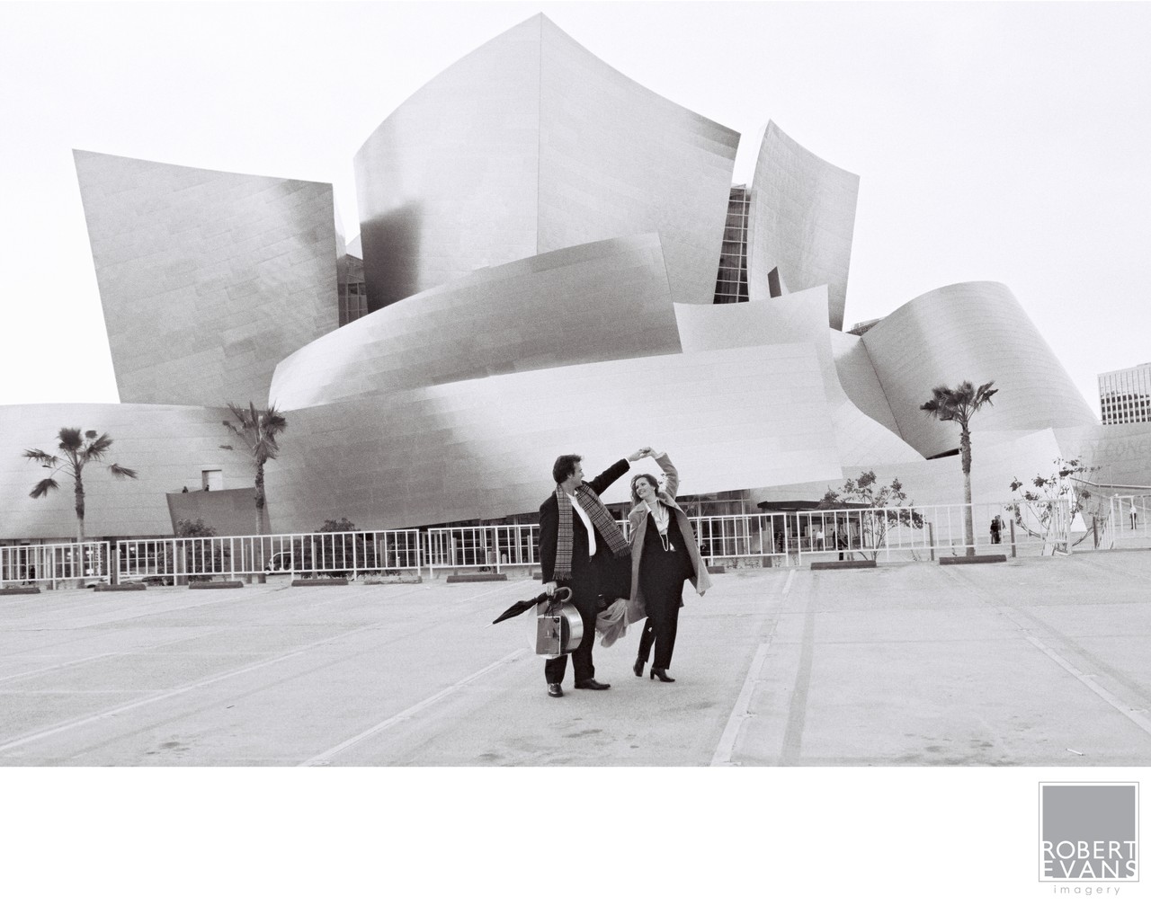 A wedding couple walks across a plaza beneath sweeping metal architecture in black and white.