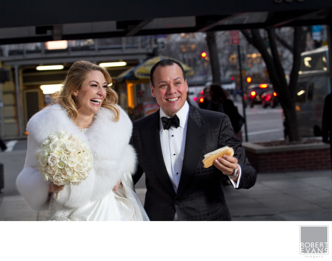 A bride in a white wrap laughs beside her groom as he holds a snack outdoors.