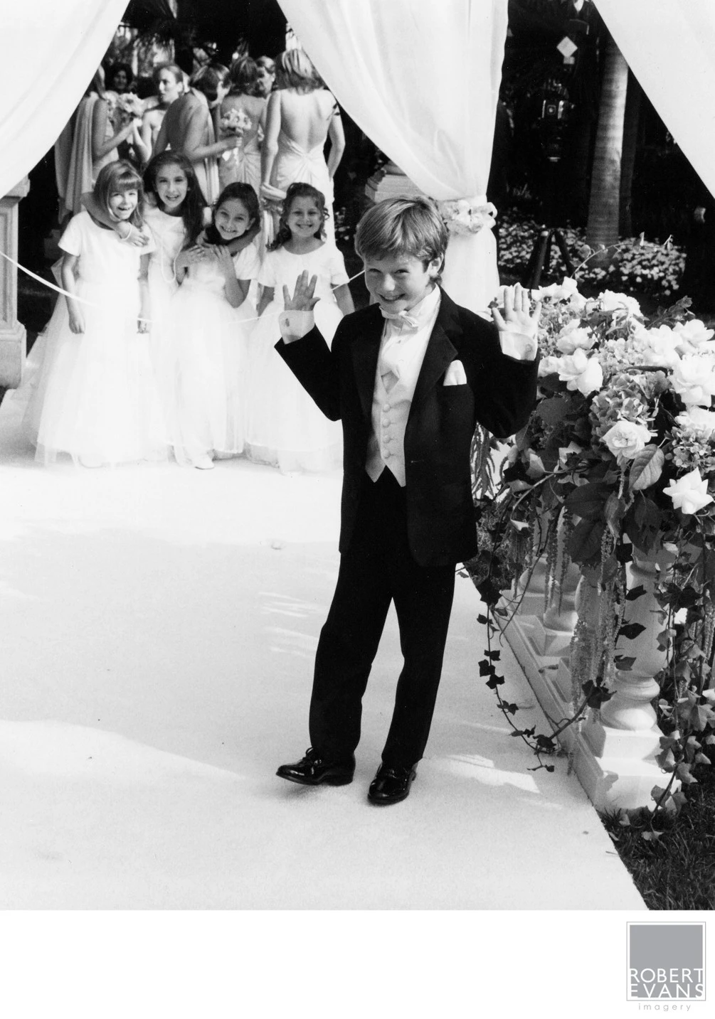 A small boy in formal clothing walks ahead of a bride and wedding attendants in black and white.