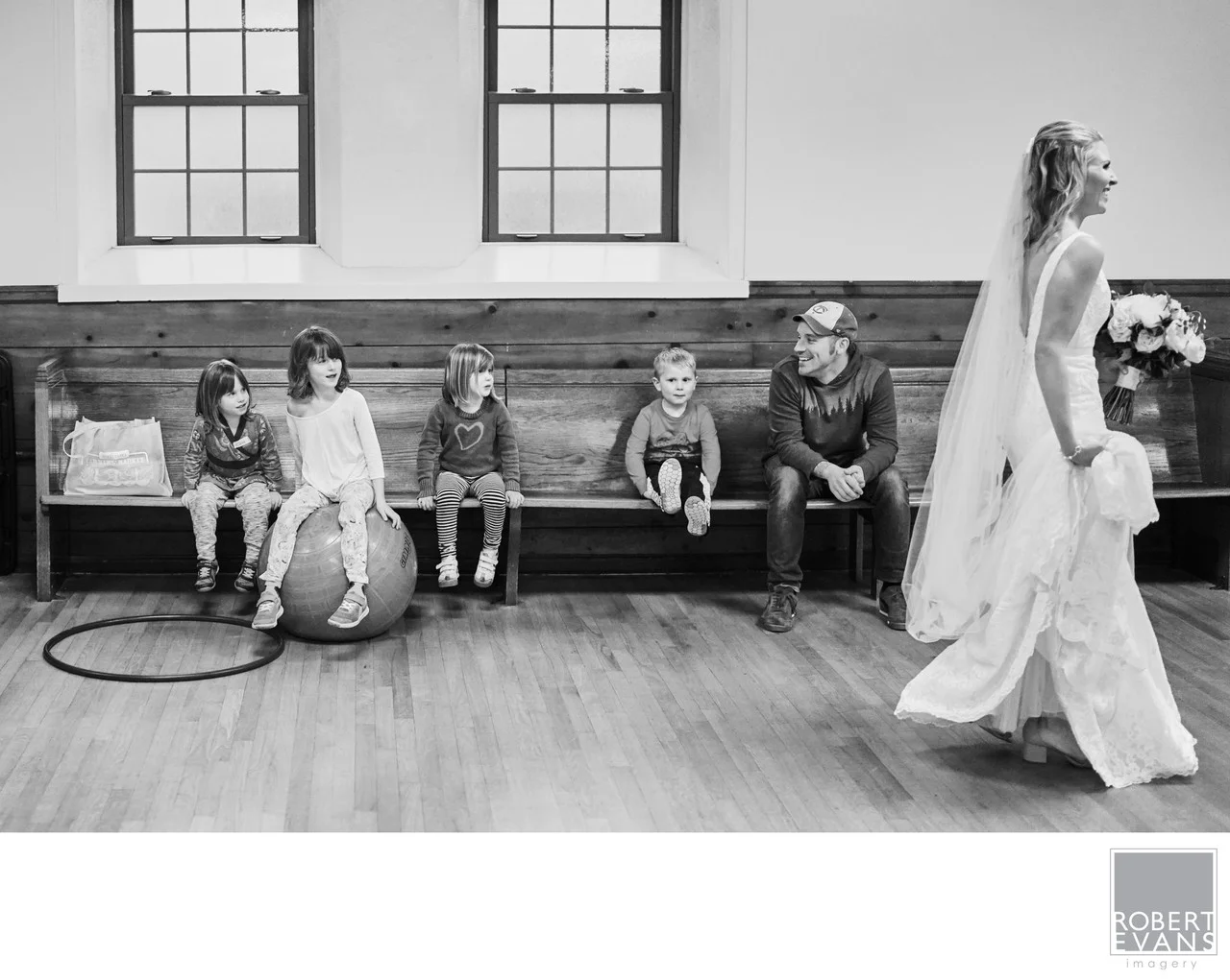 Children sit along a church bench watching a bride walk past in black and white.