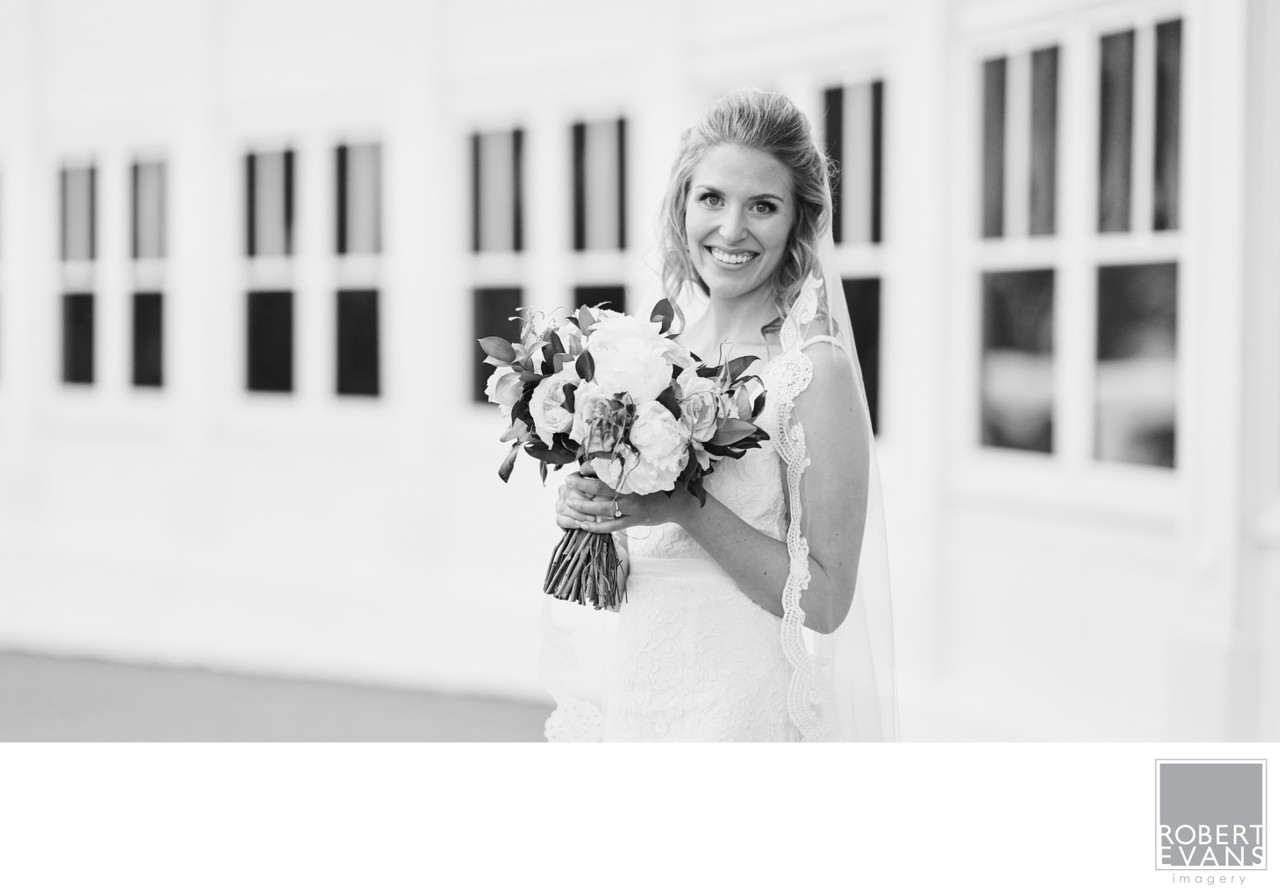 A bride holds her bouquet and smiles beside a wall of framed pictures in black and white.