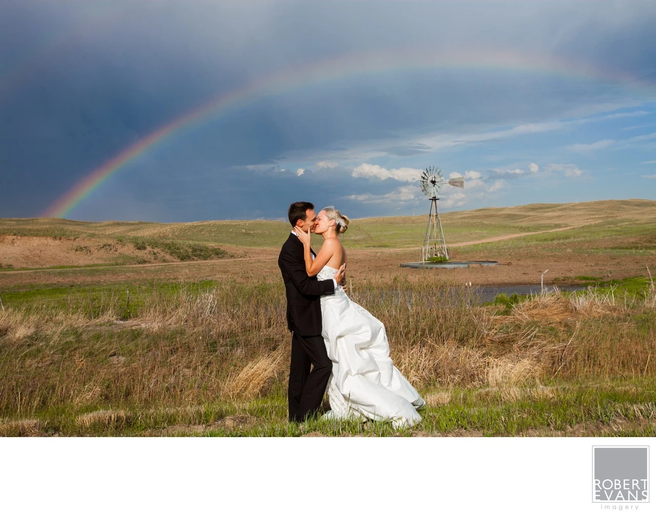 A bride and groom kiss in open countryside beneath a rainbow and dark clouds.