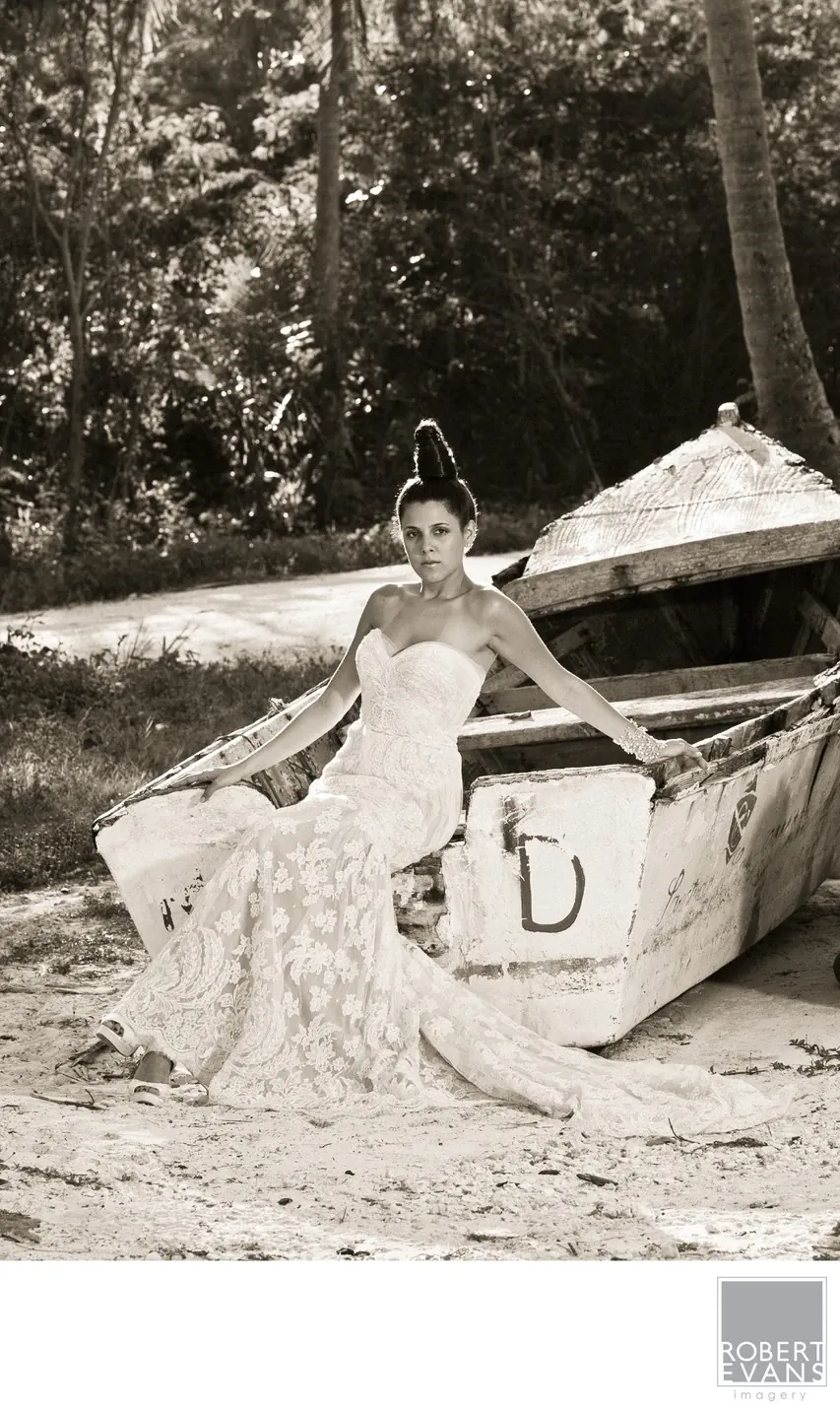 A bride sits beside a canoe at the edge of a wooded shoreline in a monochrome portrait.