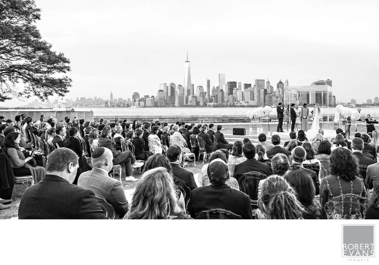 Guests watch an outdoor wedding ceremony beside the water with the New York skyline behind it.