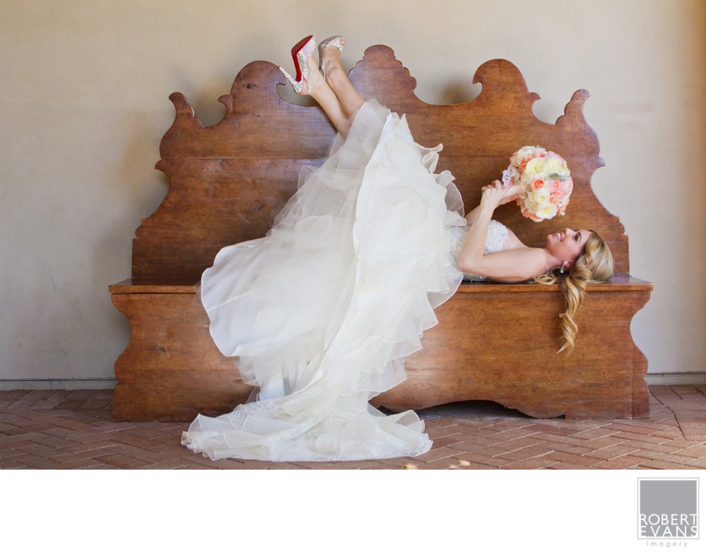 A bride reclines playfully on a carved wooden bench holding a colorful bouquet.