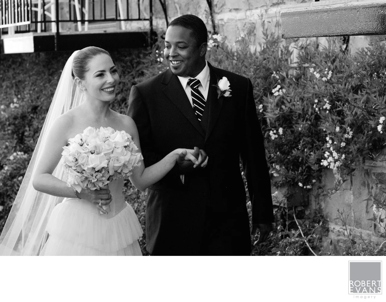 A smiling bride and groom stand together beside a weathered wall in black and white.