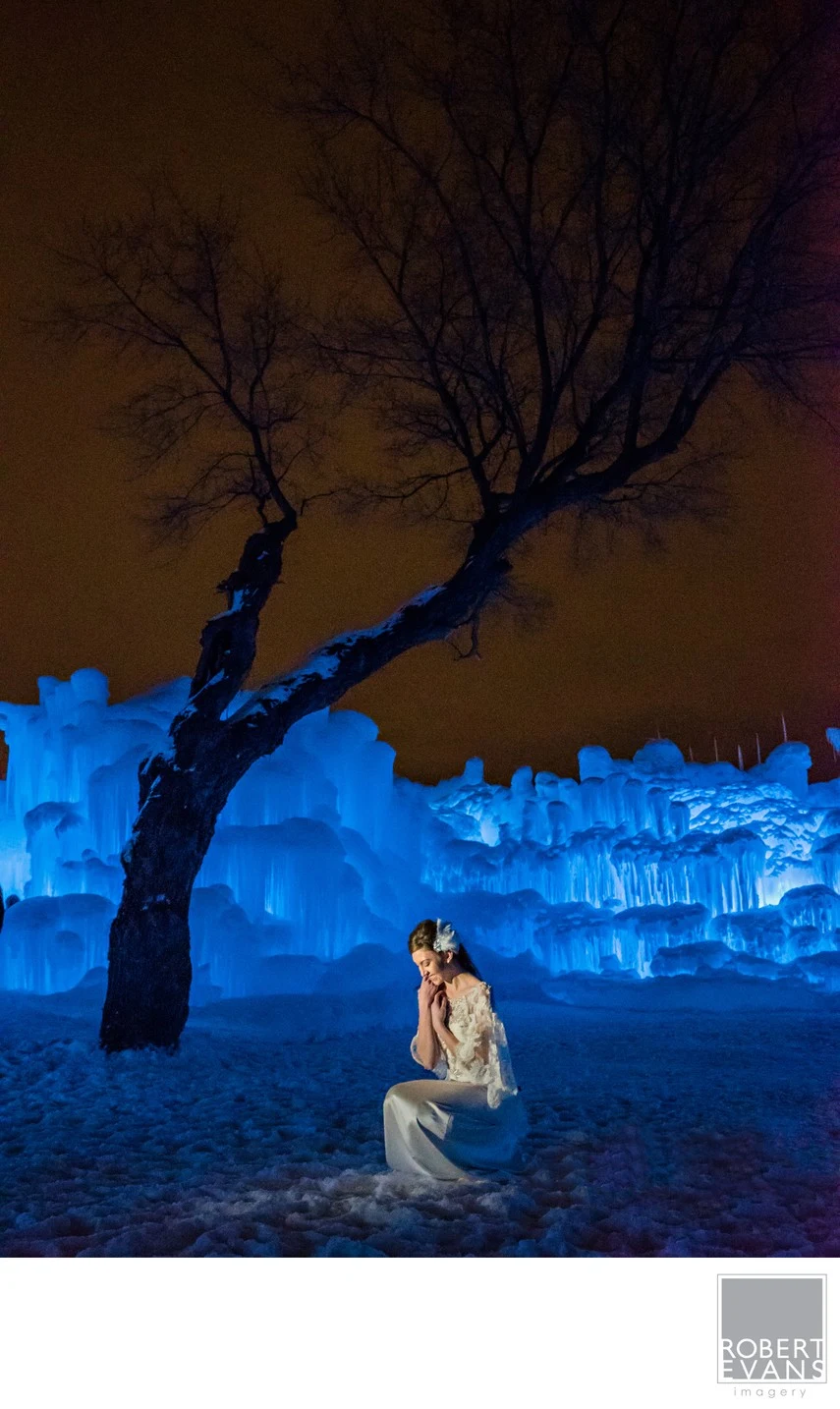 A bride sits beneath a bare tree beside blue-lit ice formations at night.