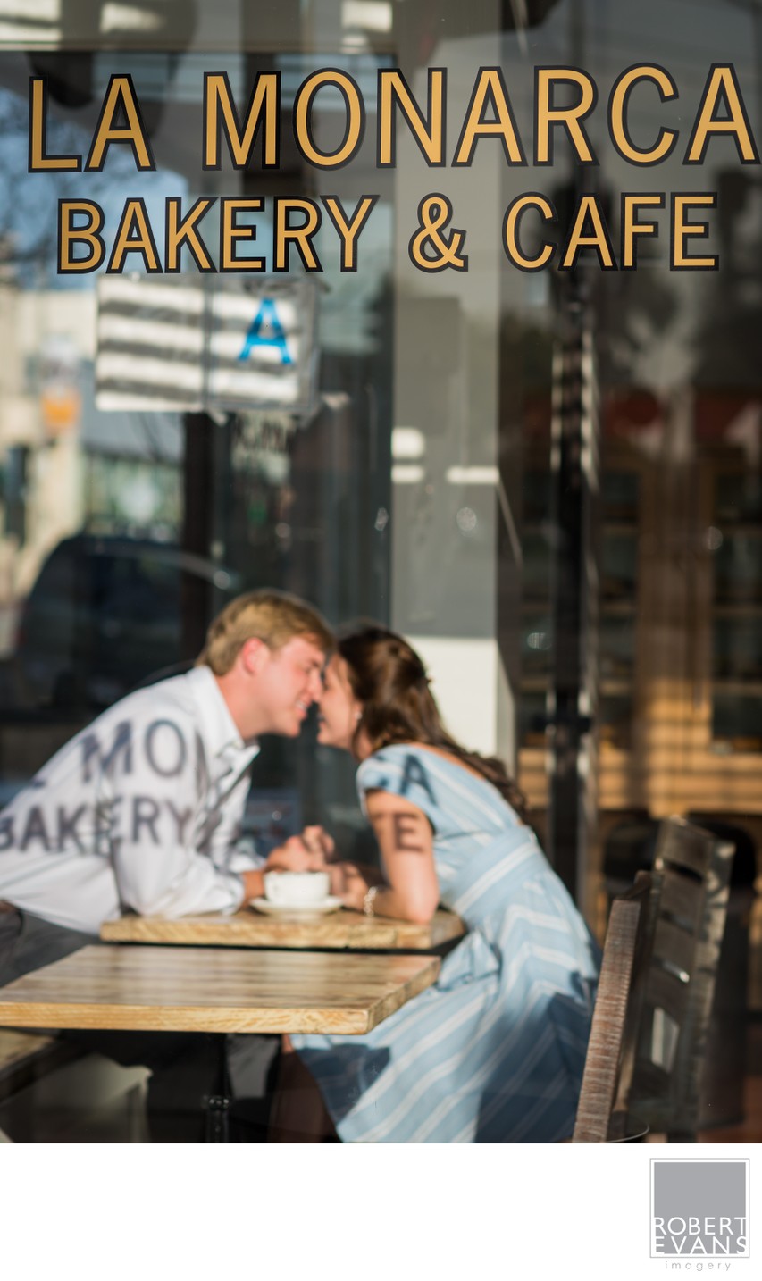 A couple leans together at a café table seen through a storefront window.