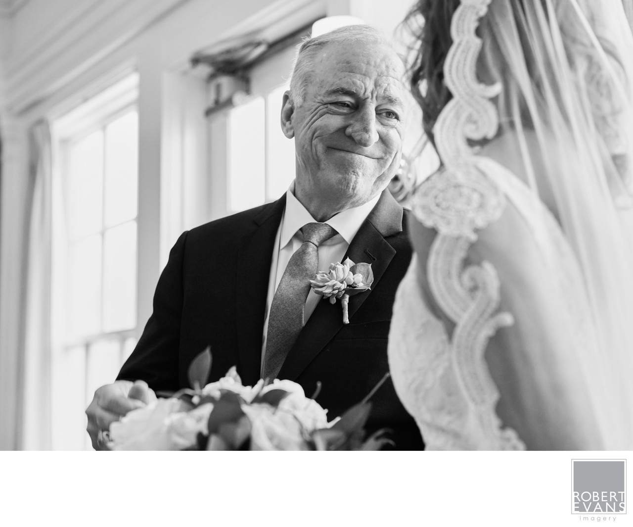 An older man smiles warmly toward a veiled bride in a close black-and-white photograph.