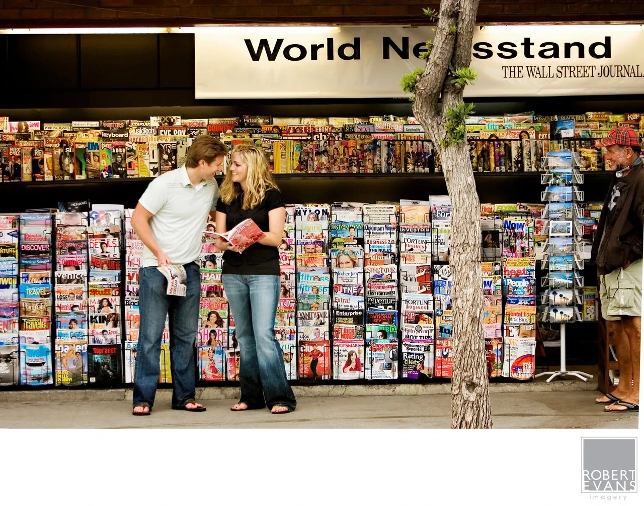 A couple leans together in front of a newsstand covered with colorful magazines.