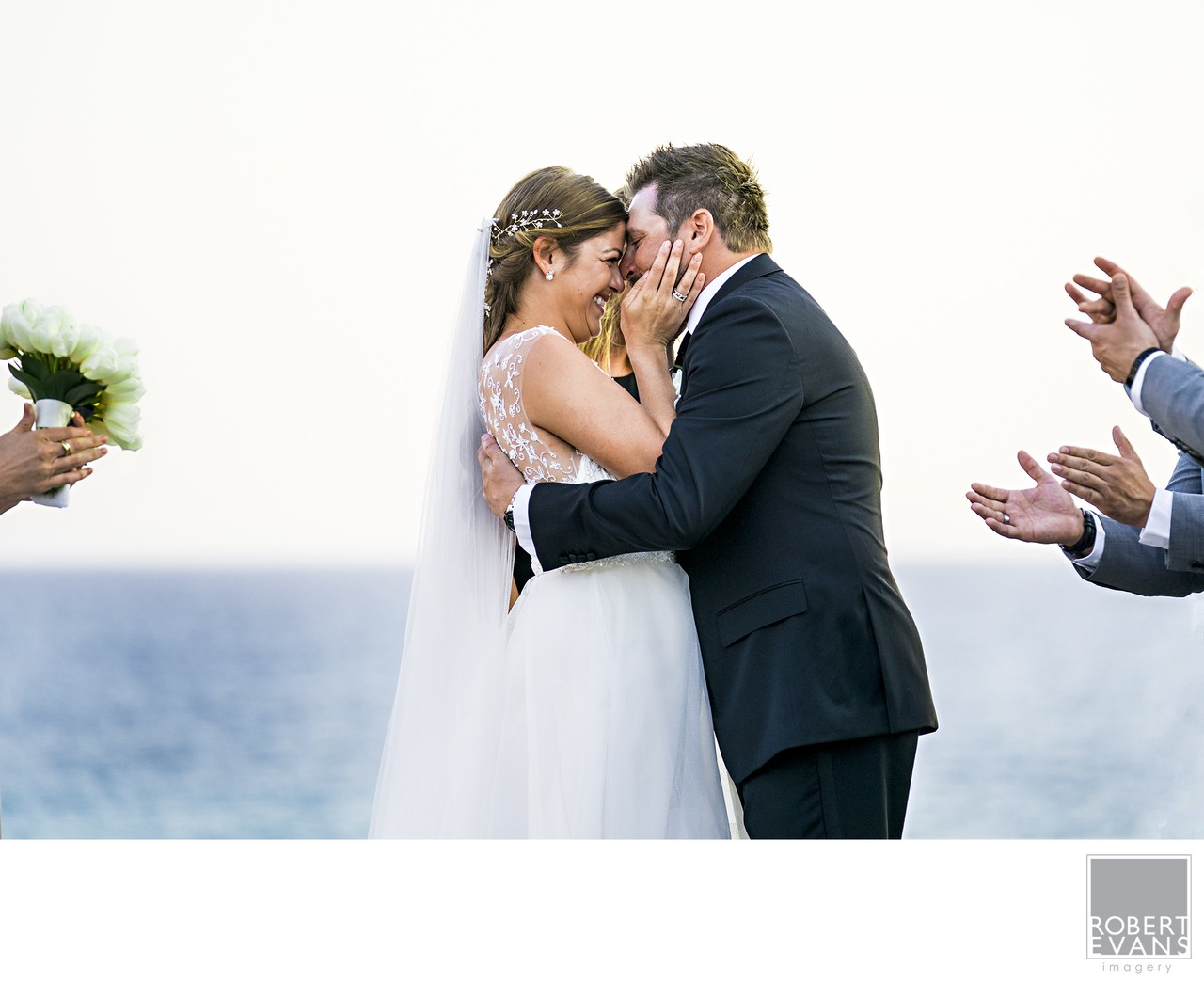 A bride and groom kiss beside the ocean while nearby guests applaud.