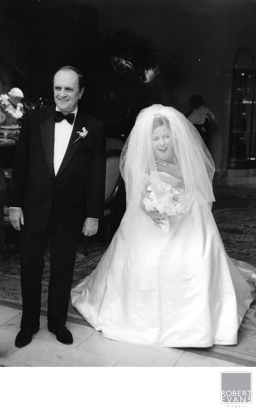 A veiled bride stands beside her father before entering the wedding ceremony in black and white.
