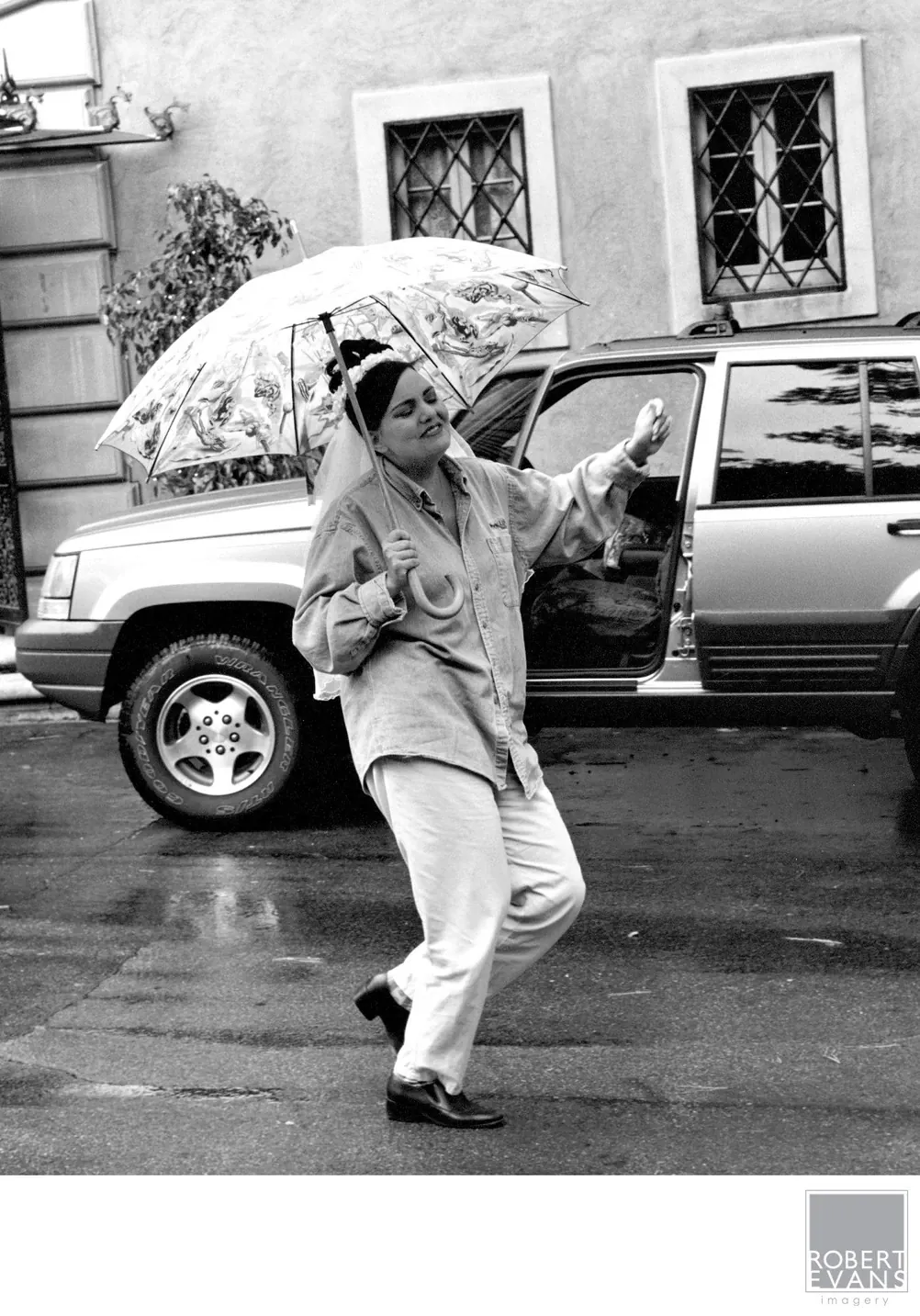 A person dances under a patterned umbrella on a wet street in black and white.