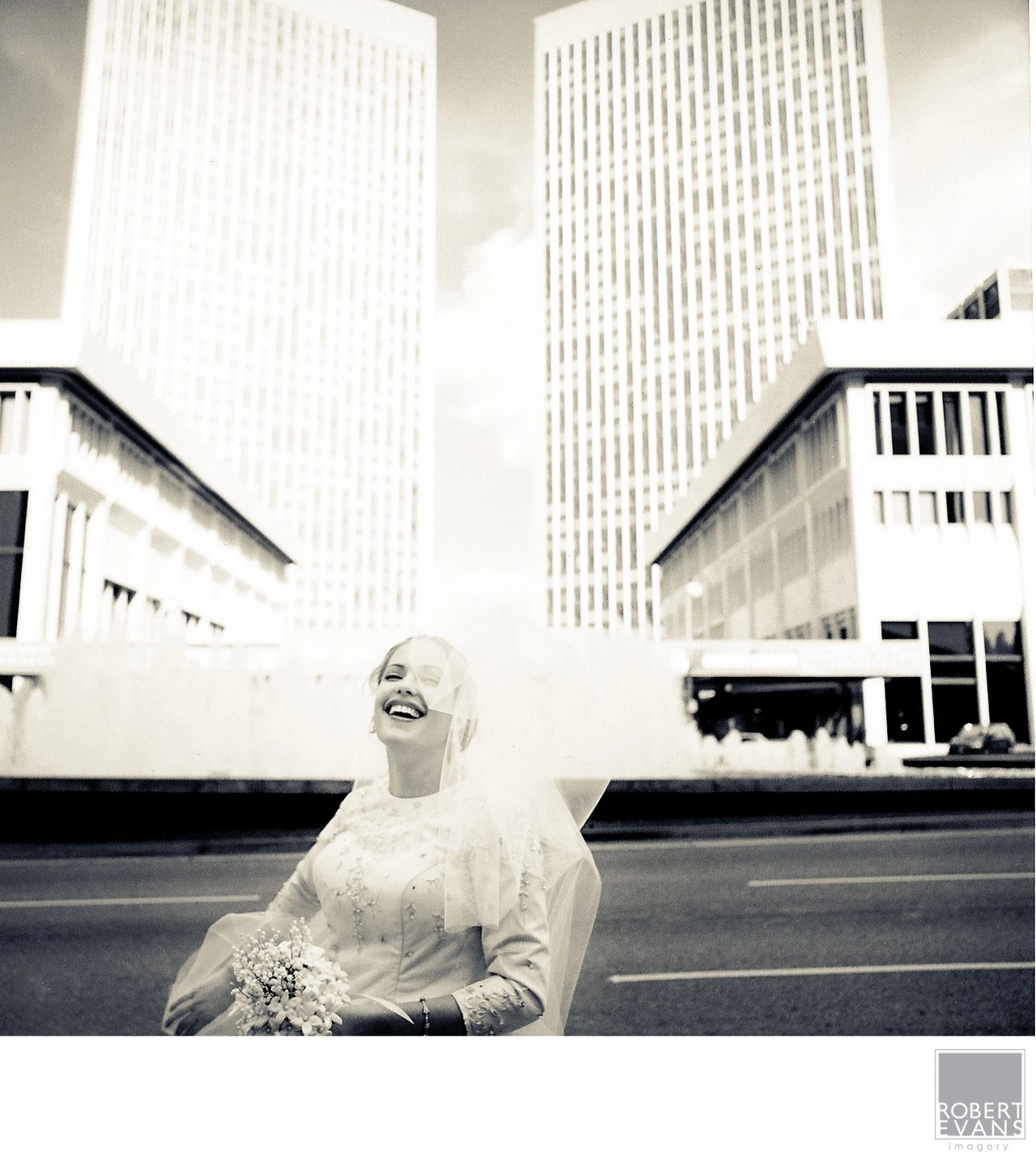 A bride laughs beneath tall downtown buildings in a candid black-and-white portrait.