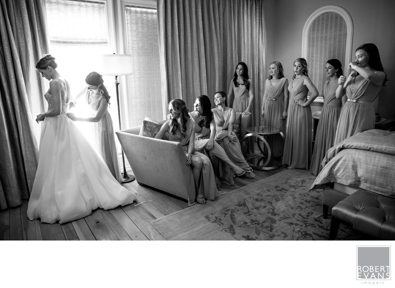 A bride stands near a window while bridesmaids wait around a softly lit room in black and white.