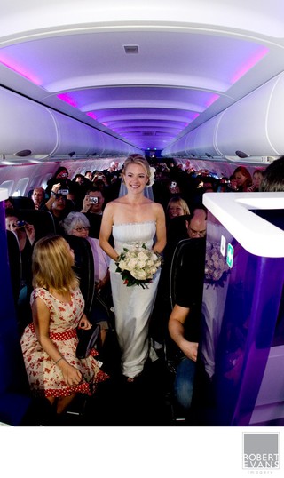 A bride holding a bouquet stands among passengers inside an airplane cabin with purple lighting.