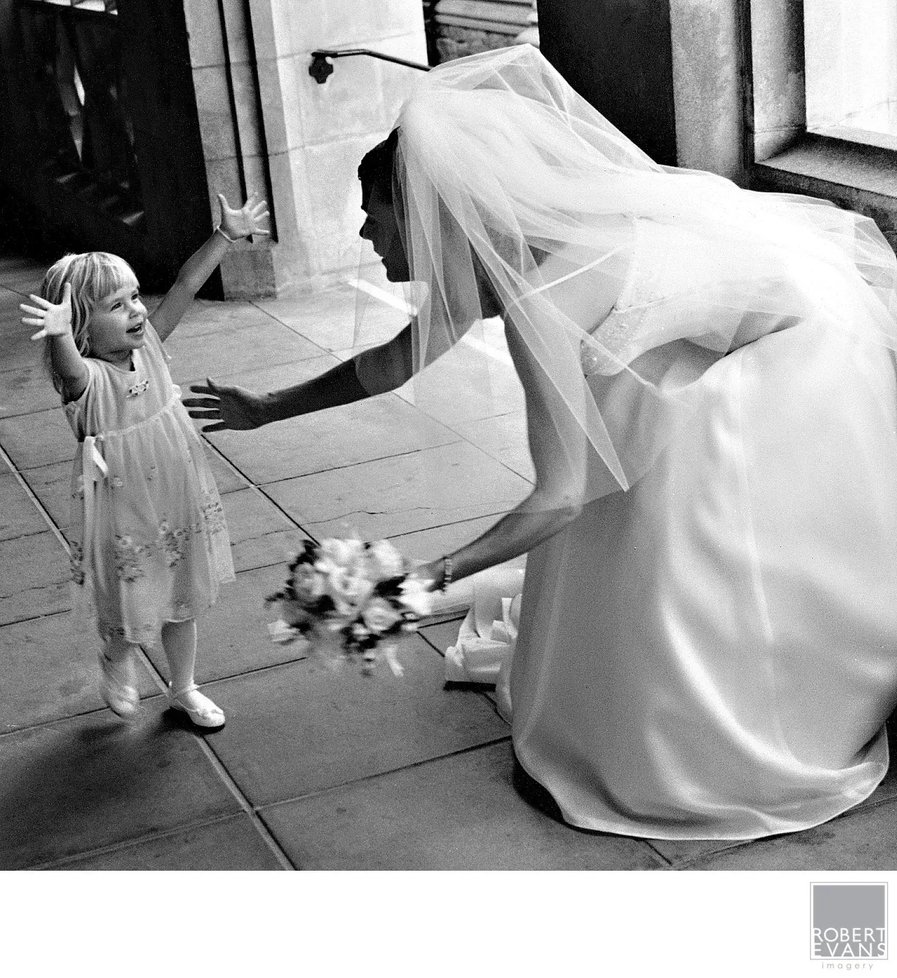 A bride bends toward a small child outside a building in black and white.