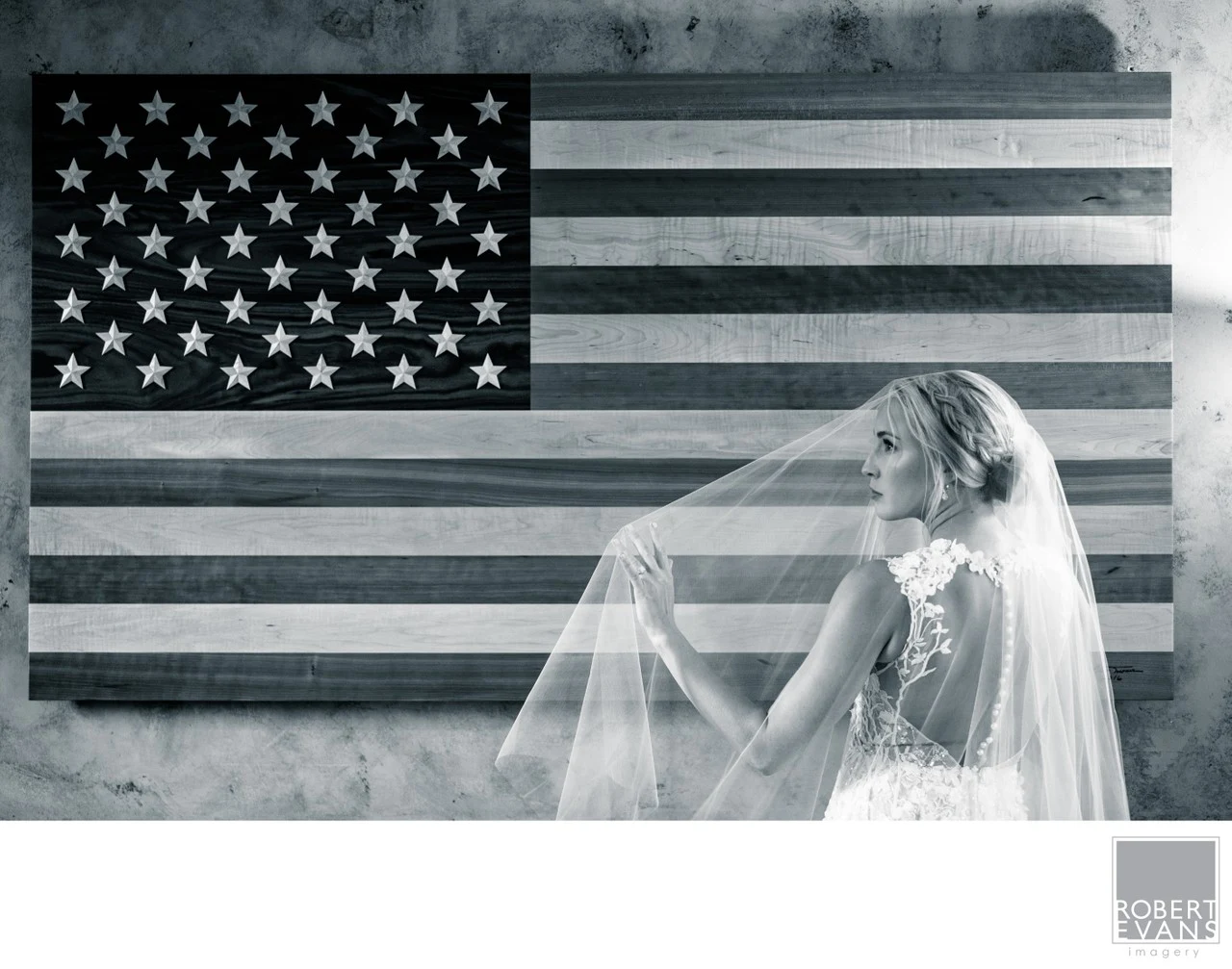 A bride lifts her veil beneath a large American flag in a black-and-white portrait.