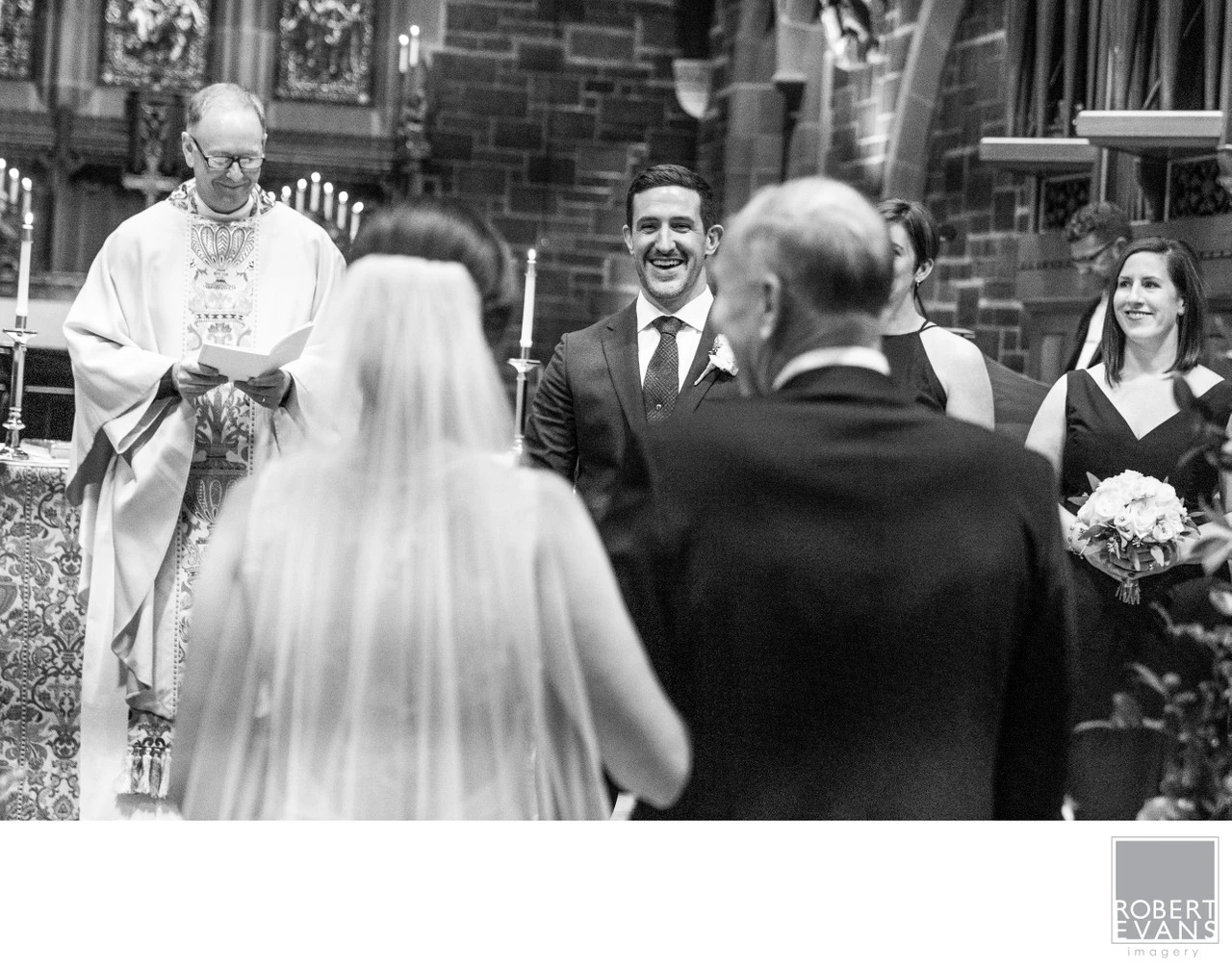 A smiling groom faces his bride and officiant during a church wedding in black and white.